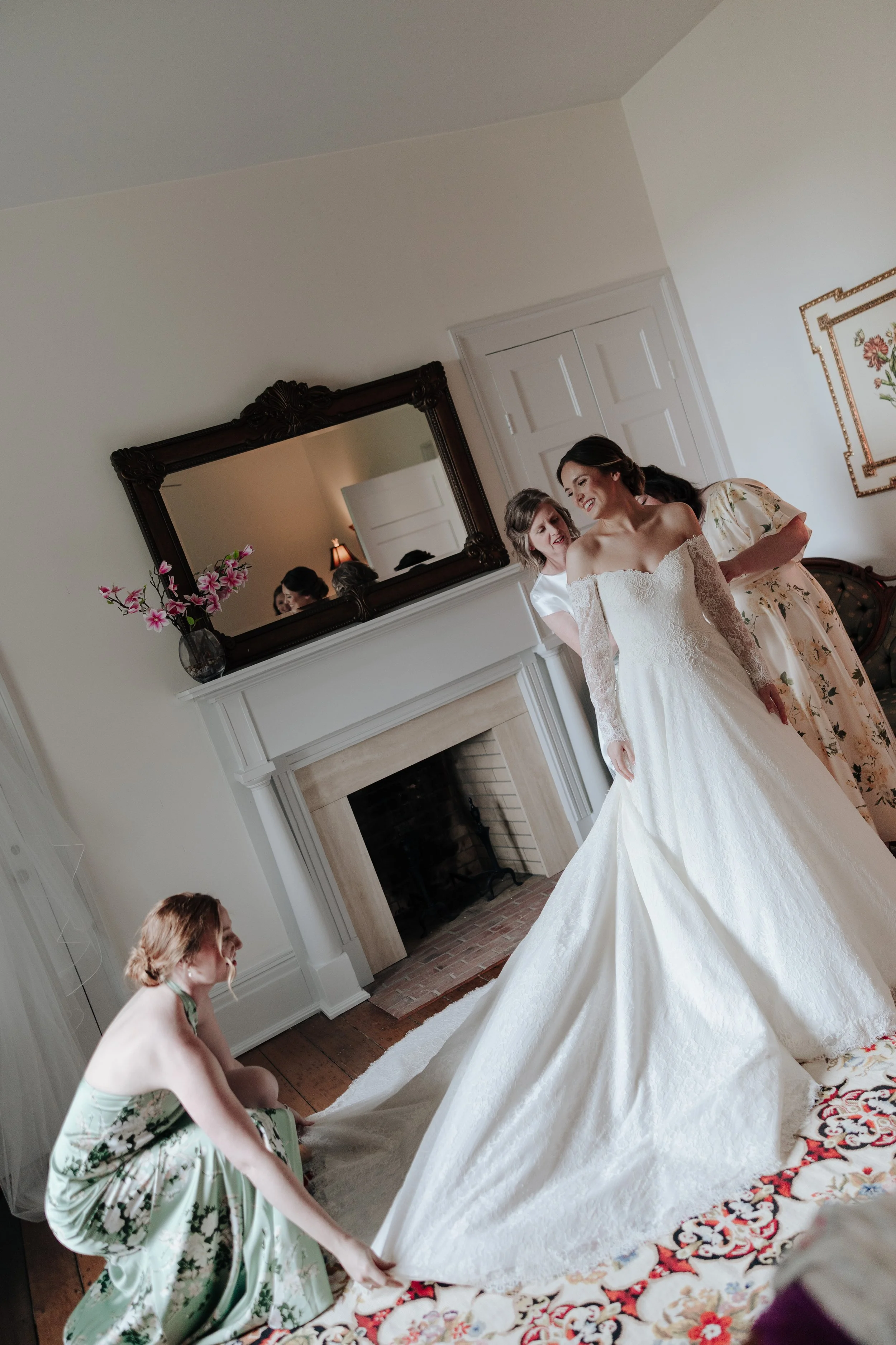 A bride in a white wedding dress is being assisted by four women, one in a floral dress and one in a white blouse, in a spacious room with a fireplace, a large mirror, and a vase of pink flowers on the mantel.
