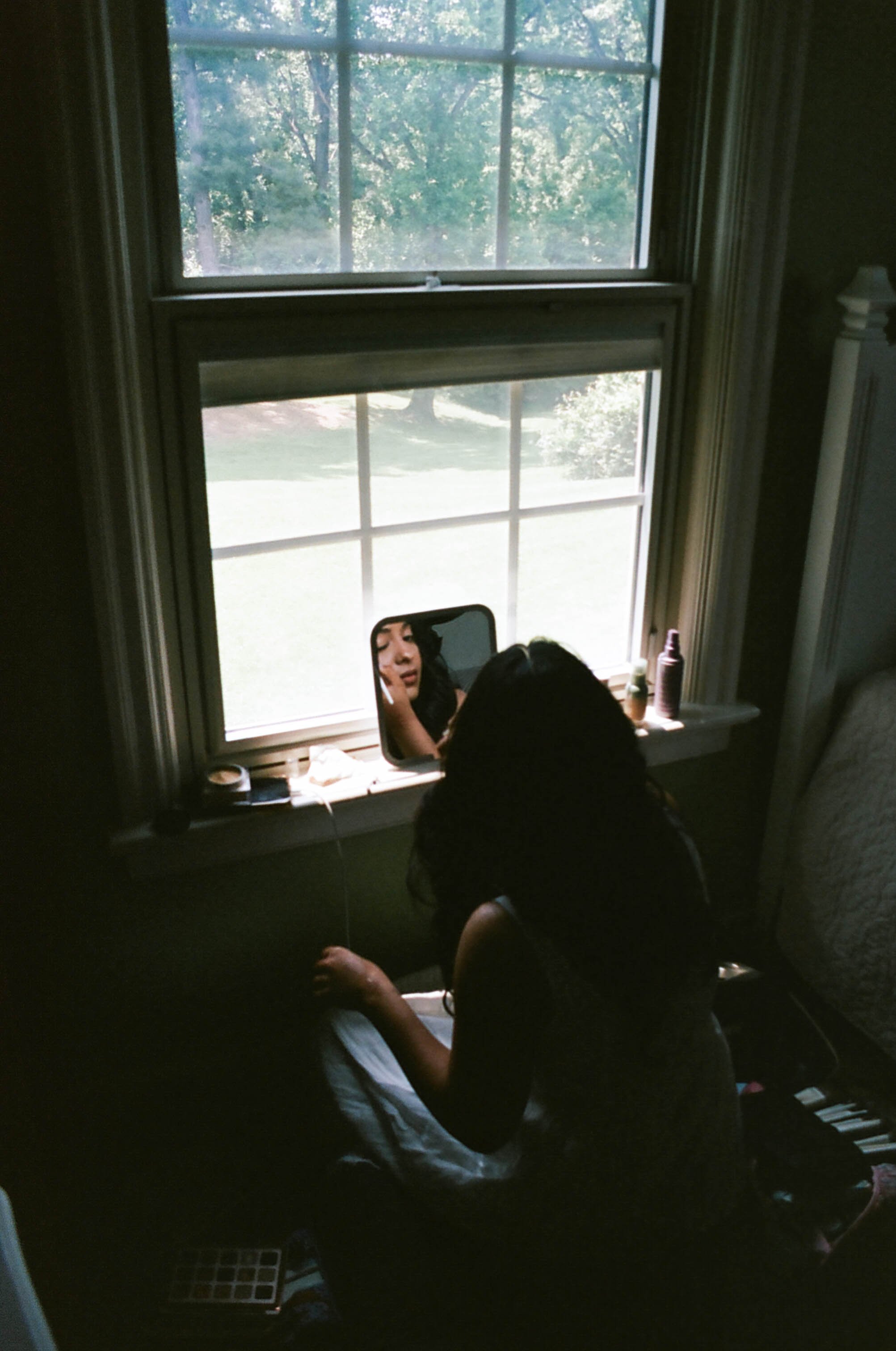 A person is sitting in front of a window, reflected in a small mirror placed on the window sill, with a second person visible in the reflection applying makeup or grooming, while outside the window, a green outdoor scene is visible.