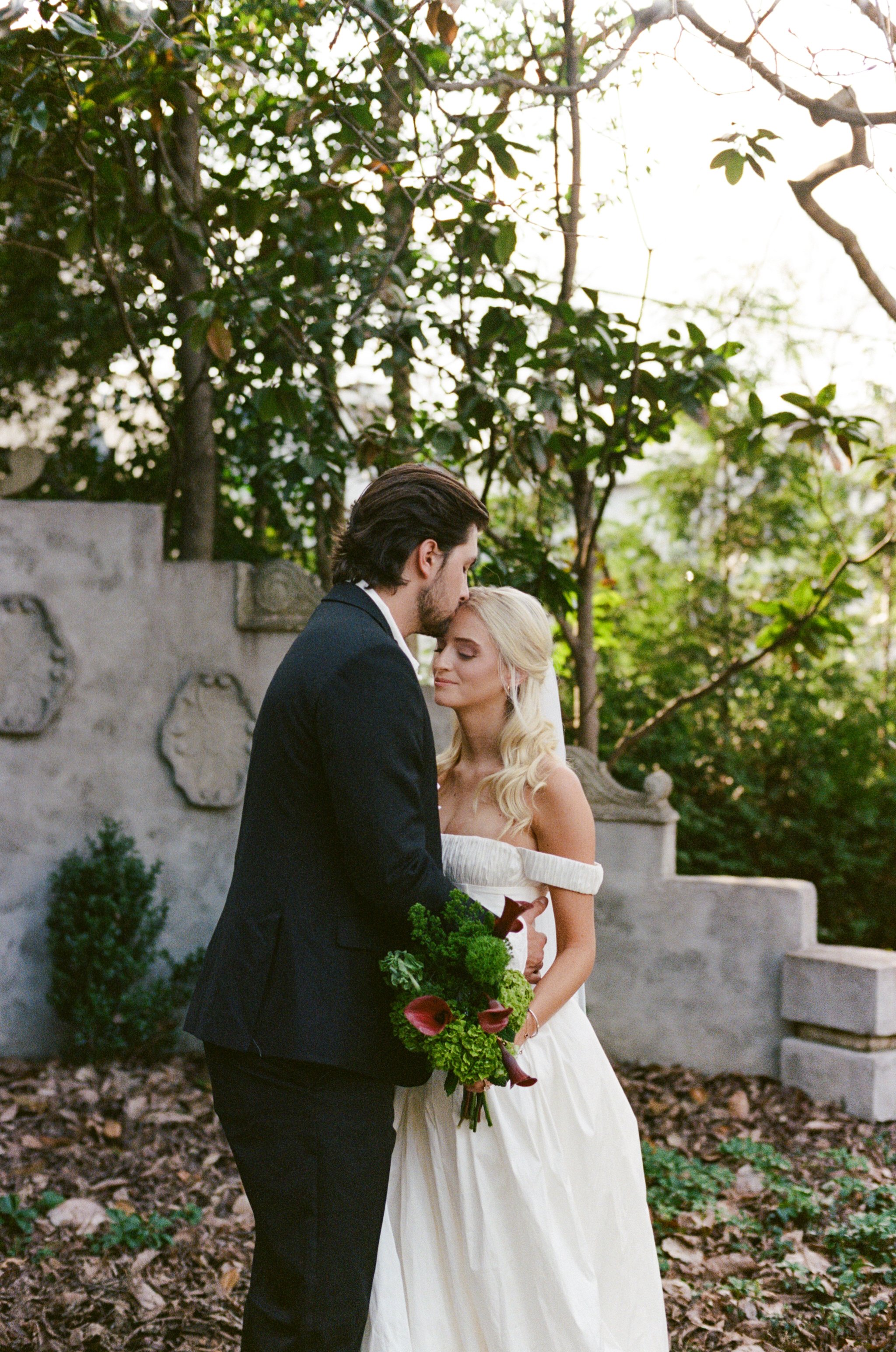 A bride and groom in wedding attire standing close together outdoors, with the groom kissing the bride's forehead. The bride holds a bouquet, and there are trees and stone structures in the background.