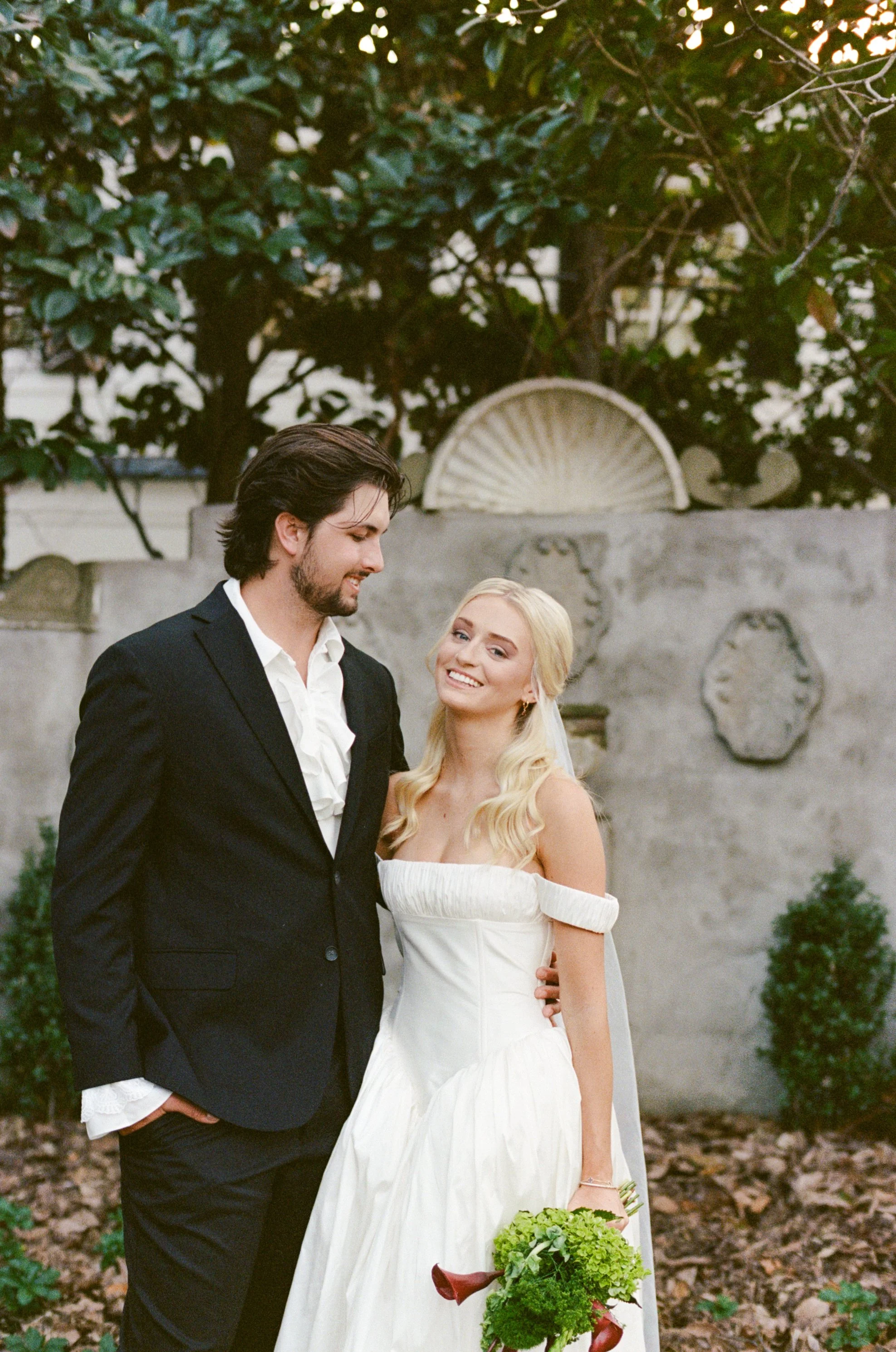 A bride and groom standing outdoors, smiling and looking at each other, with trees and a decorative wall in the background.