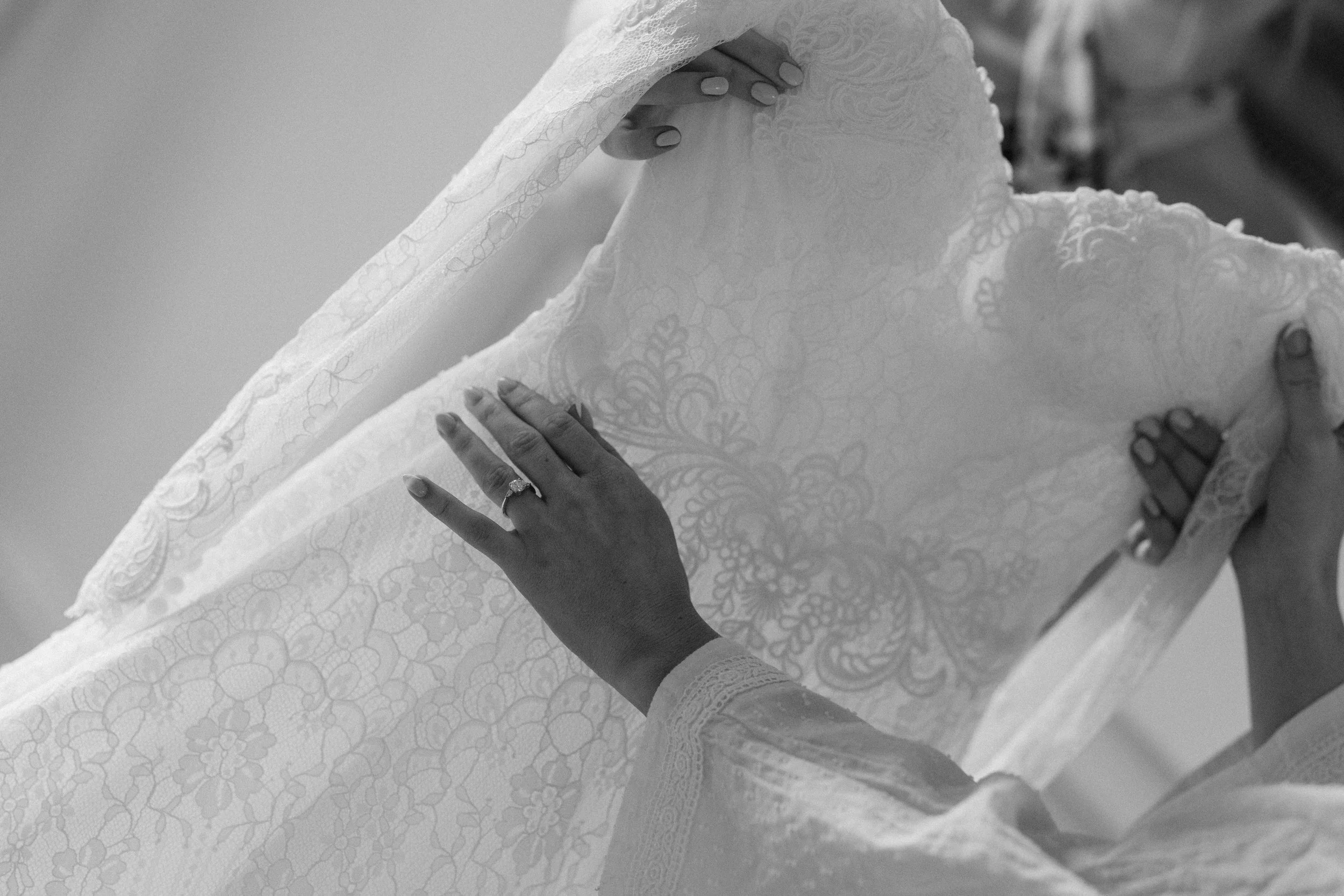 Close-up of a woman adjusting the lace veil of a wedding dress, with her hand visible wearing an engagement ring and manicured nails.