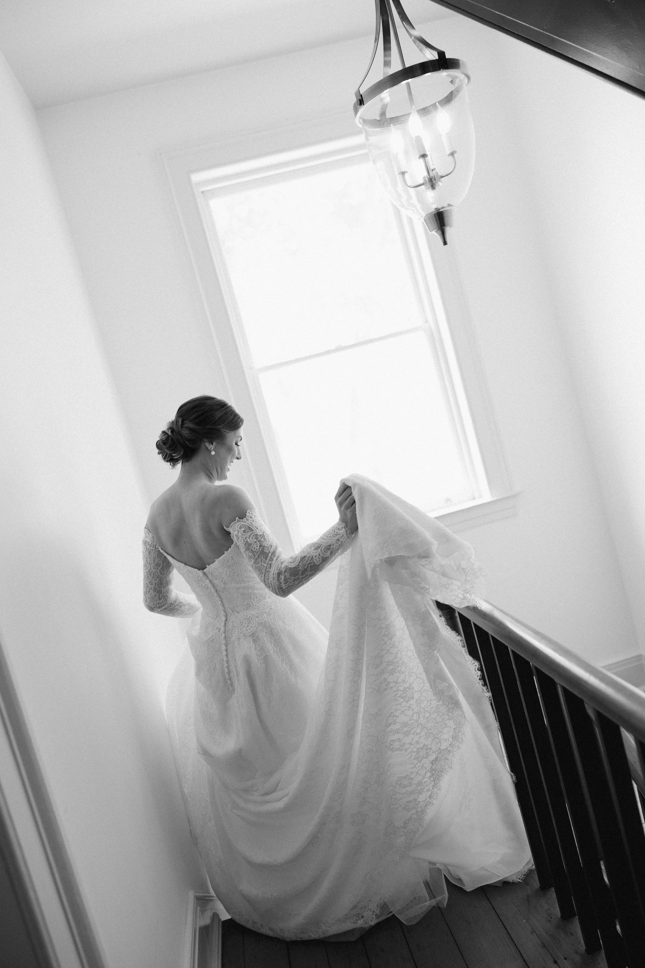 A woman in a wedding dress standing next to a staircase with a railing. She is holding the train of her dress and smiling, with natural light coming in from a window behind her.
