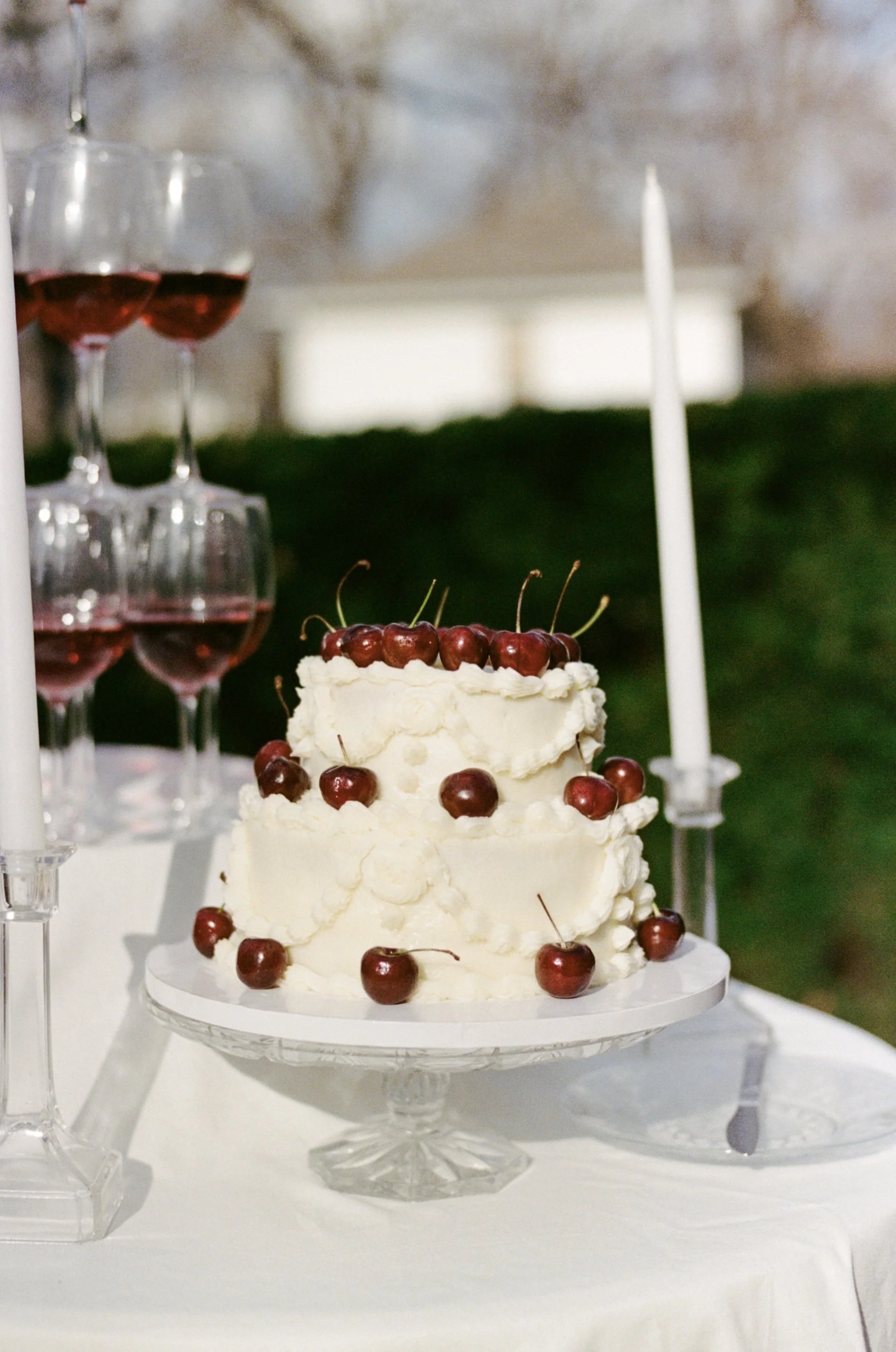 Three-tiered wedding cake topped with cherries, decorated with white frosting, on a glass cake stand. To the left, a pyramid of filled wine glasses, and to the right, two tall white candles in holders, on a white tablecloth.