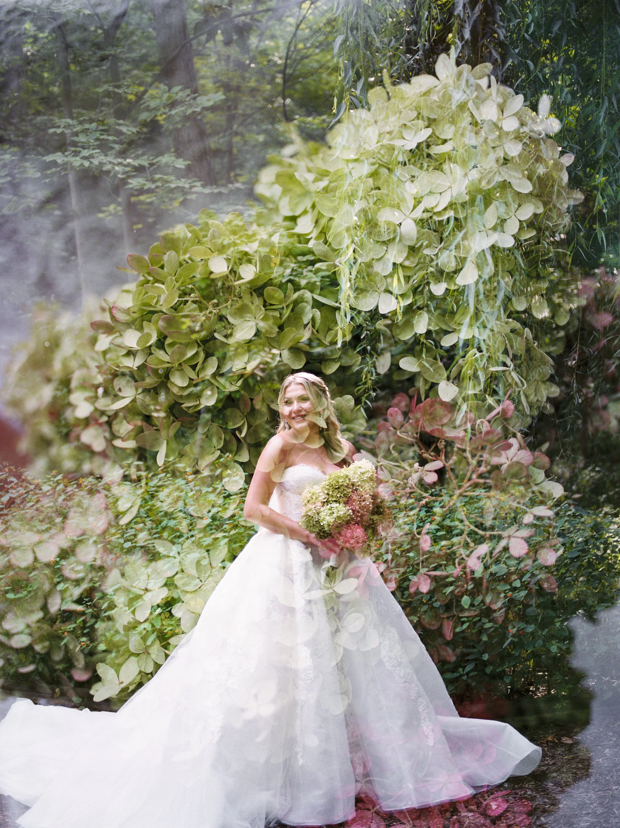 A woman in a white wedding dress holding a large bouquet of flowers, standing in front of lush green bushes and trees in a garden setting.