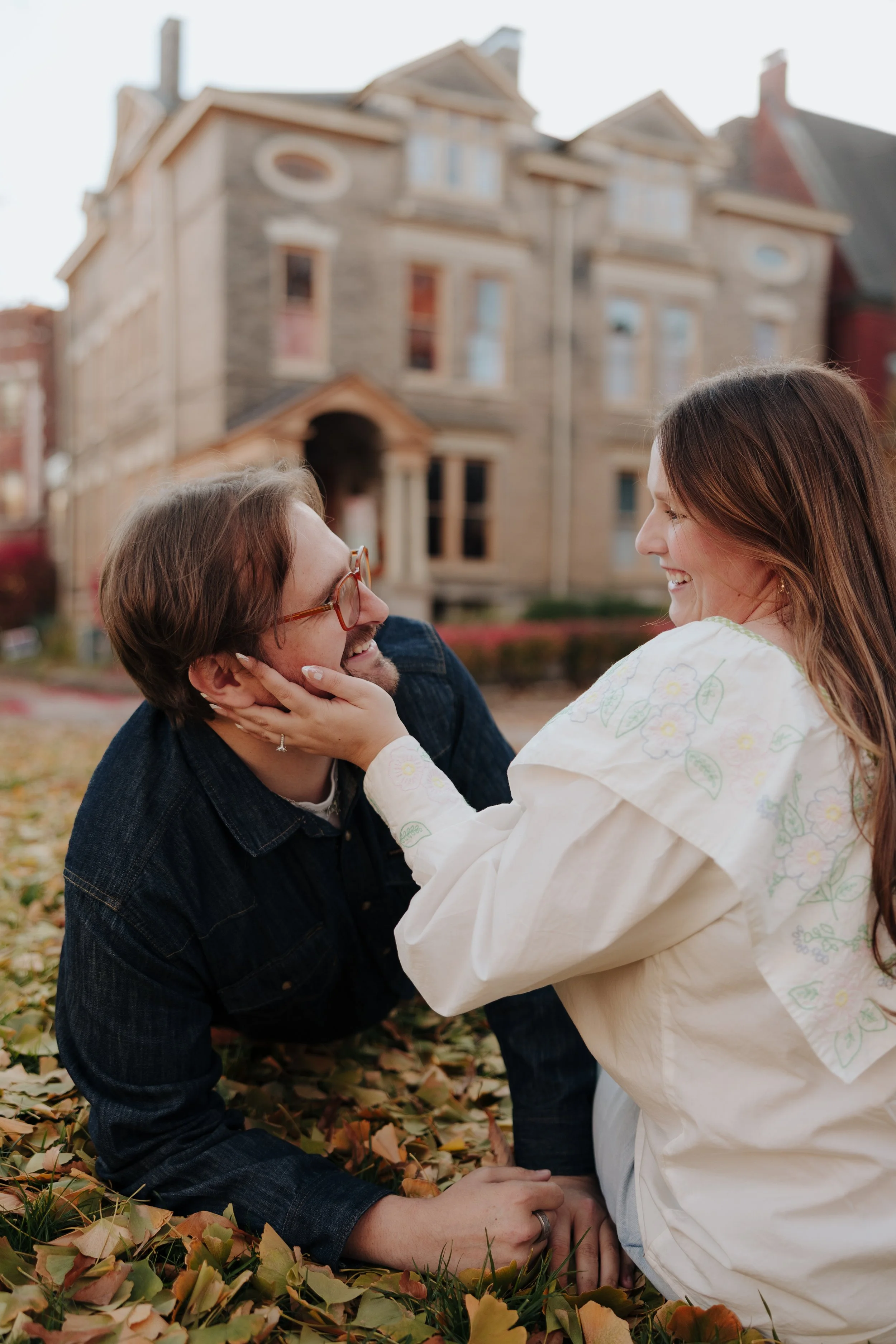 A couple playfully interacting outside on fallen autumn leaves with a large historic house in the background.