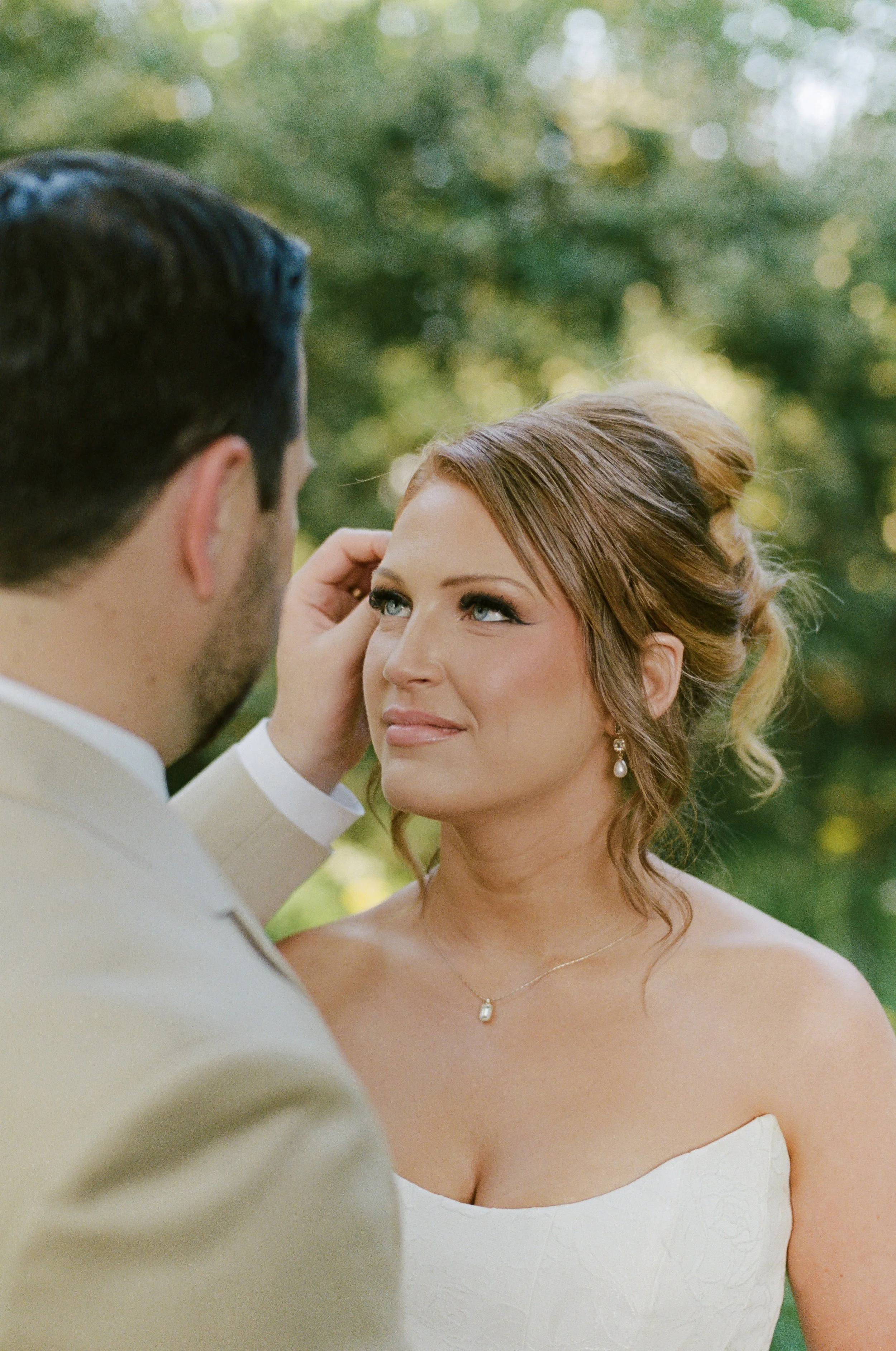 A bride and groom sharing a tender moment outdoors with a blurred background of green trees. The bride has reddish-brown hair tied back, is wearing a strapless white wedding dress, pearl earrings, and a necklace, and is looking up at the groom with a
