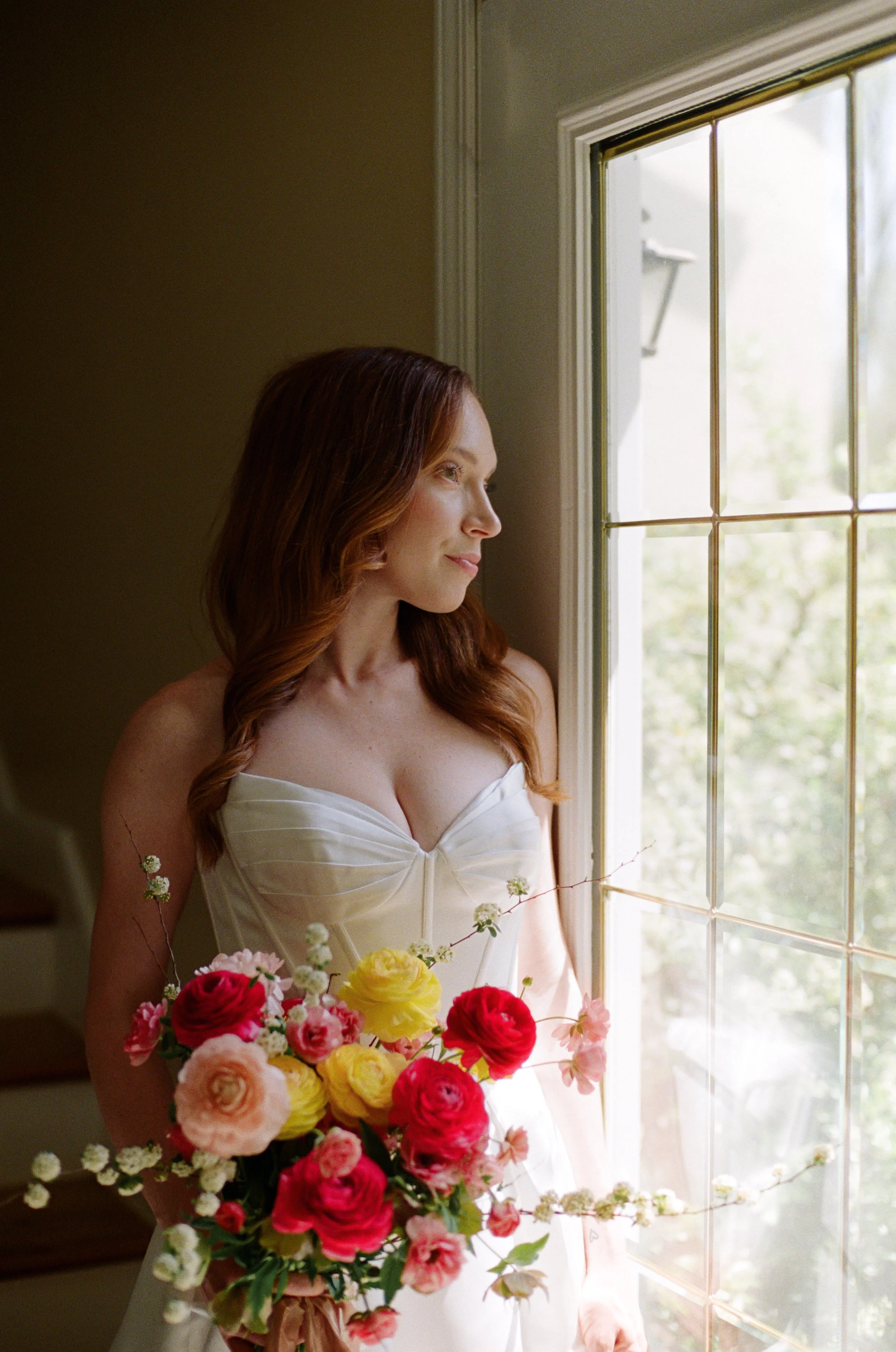 A woman with red hair wearing a cream-colored dress, holding a large bouquet of pink, red, and yellow flowers, standing by a window and looking outside.