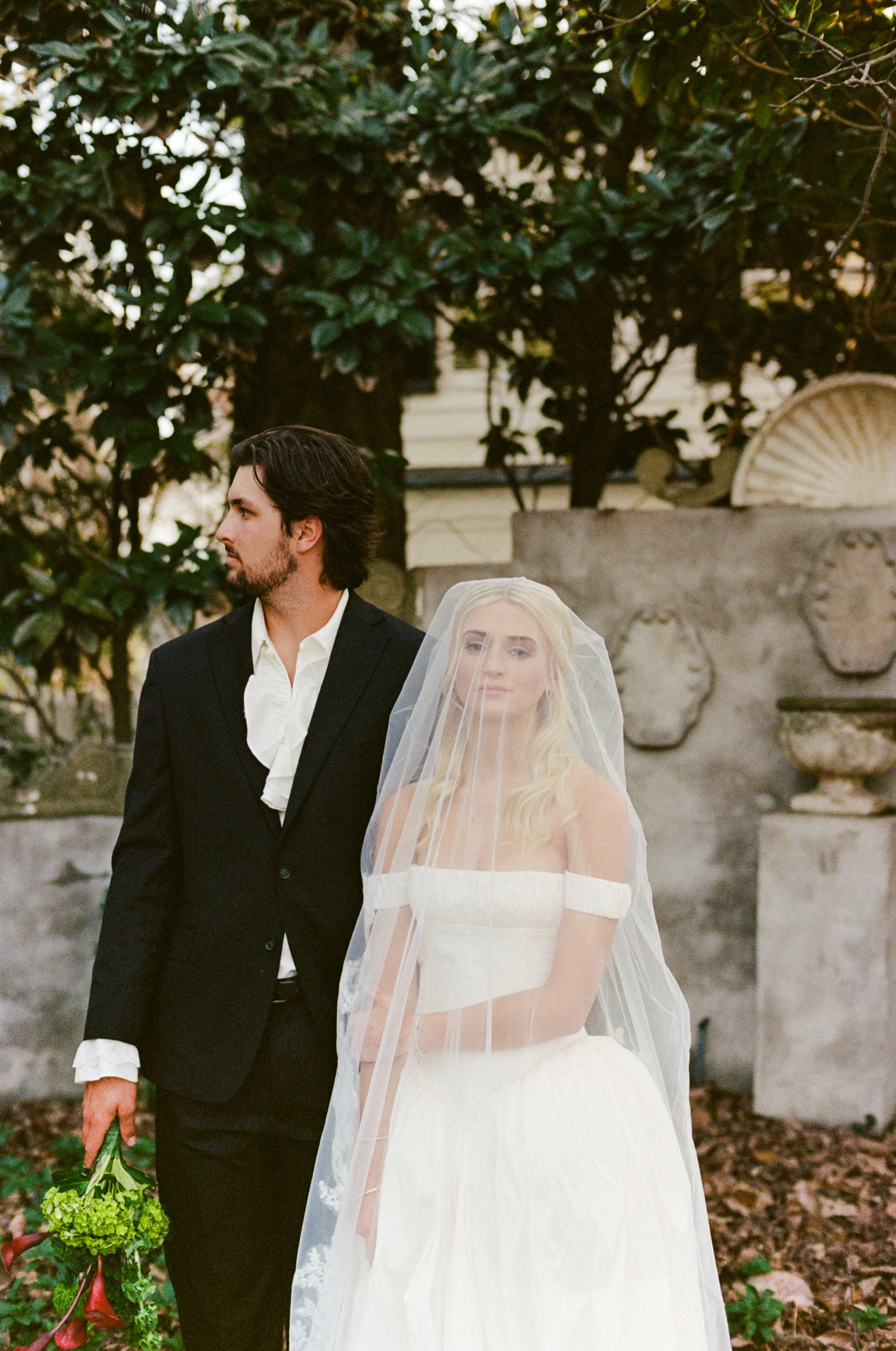 A bride and groom stand outdoors, with the bride wearing a white wedding dress and veil, and the groom dressed in a black suit holding a small bouquet of green flowers, against a background of trees and a stone wall.