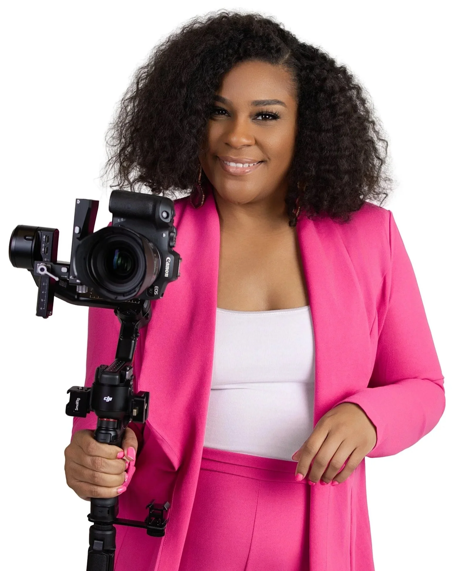 A woman with curly hair wearing a pink blazer and matching pants, holding a camera mounted on a stabilizer, smiling at the camera.
