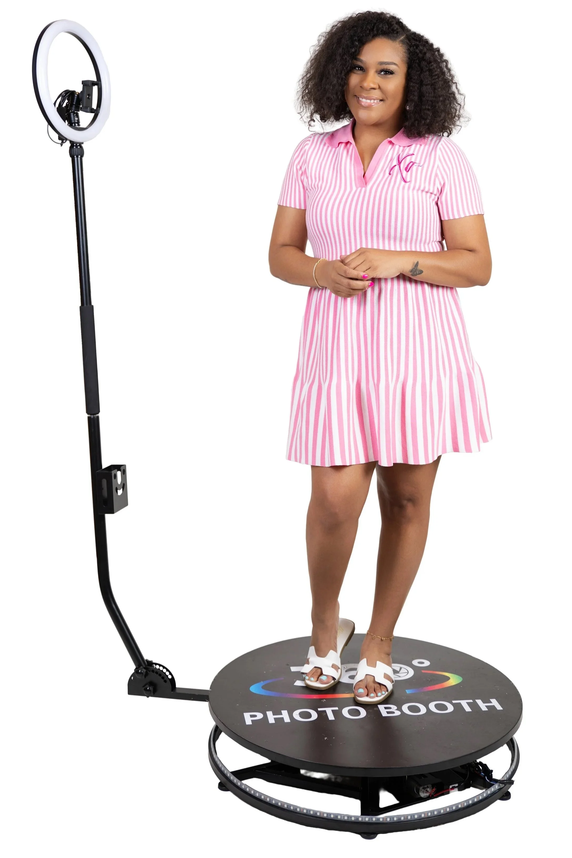 A woman in a pink and white striped dress stands on a circular photo booth platform with a ring light on a stand next to her.