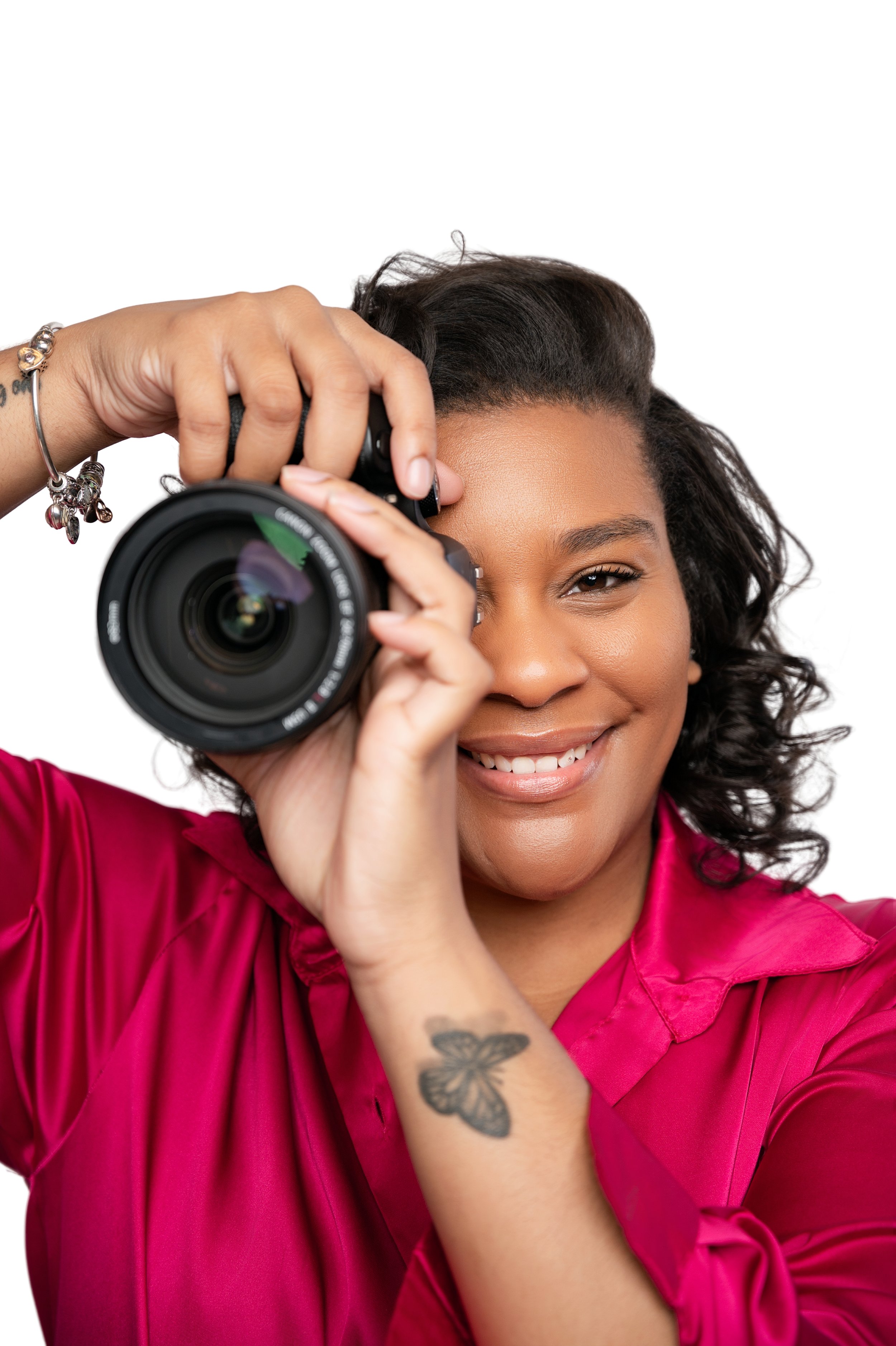 A woman with curly hair, wearing a bright pink blouse, taking a picture with a camera, smiling, showing a butterfly tattoo on her arm.