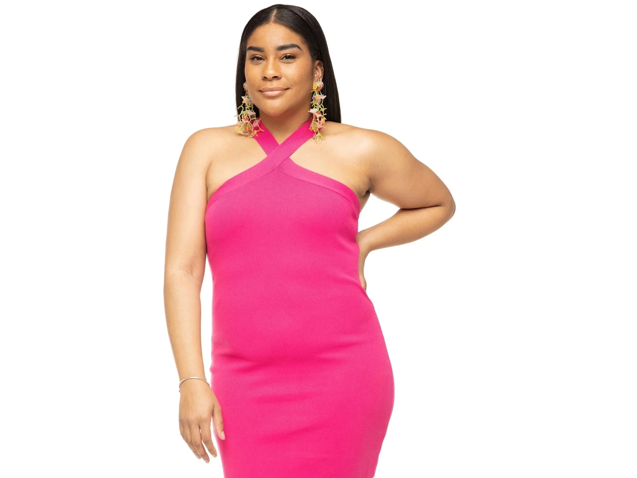 Woman wearing a pink halter dress with colorful earrings, standing with one hand on her hip, smiling softly.