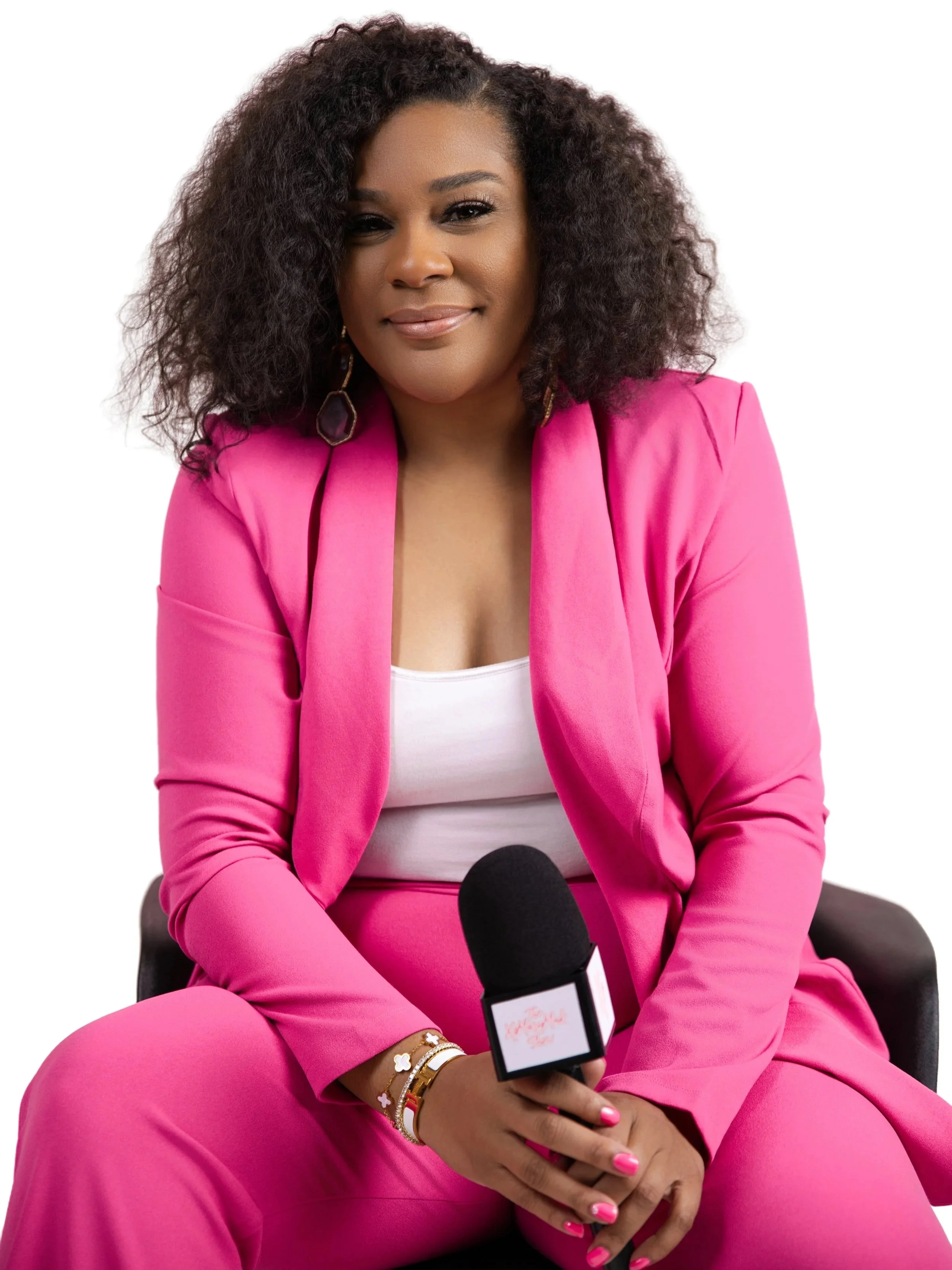 A woman wearing a bright pink suit holding a microphone and sitting on a black chair against a white background.
