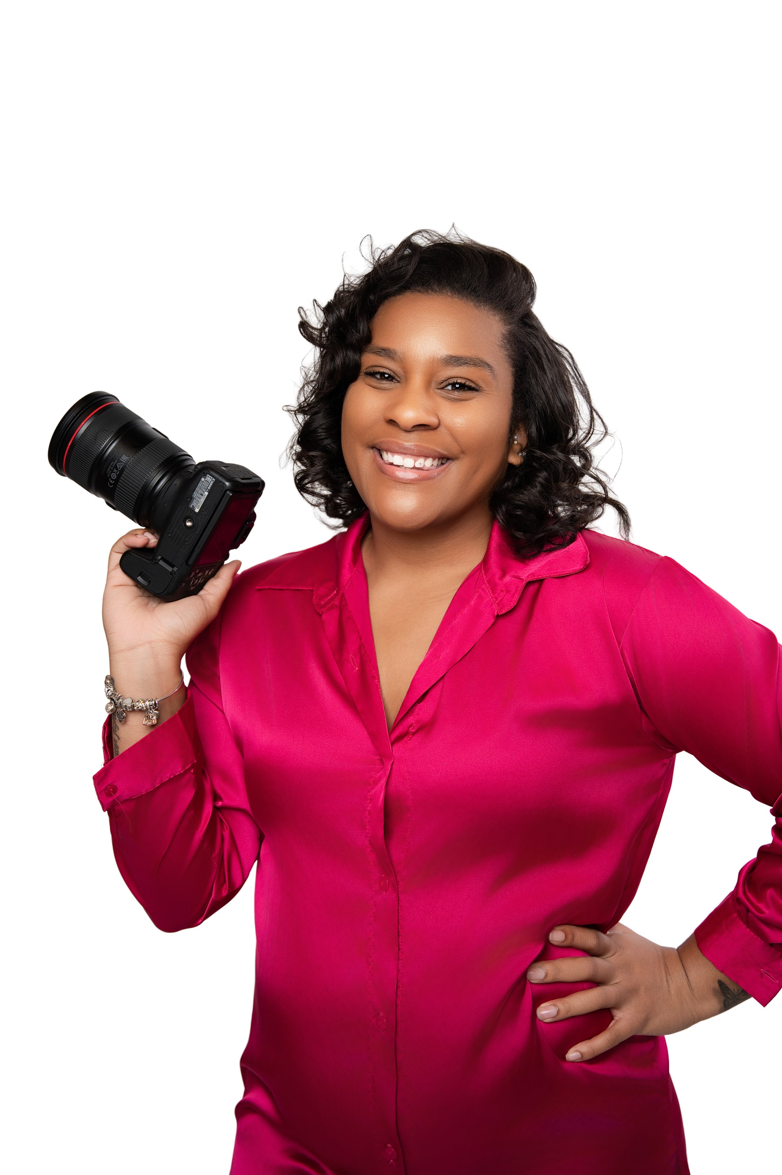 A woman with dark, curly hair smiling while holding a professional camera with a large lens, wearing a bright pink satin blouse, standing against a white background.