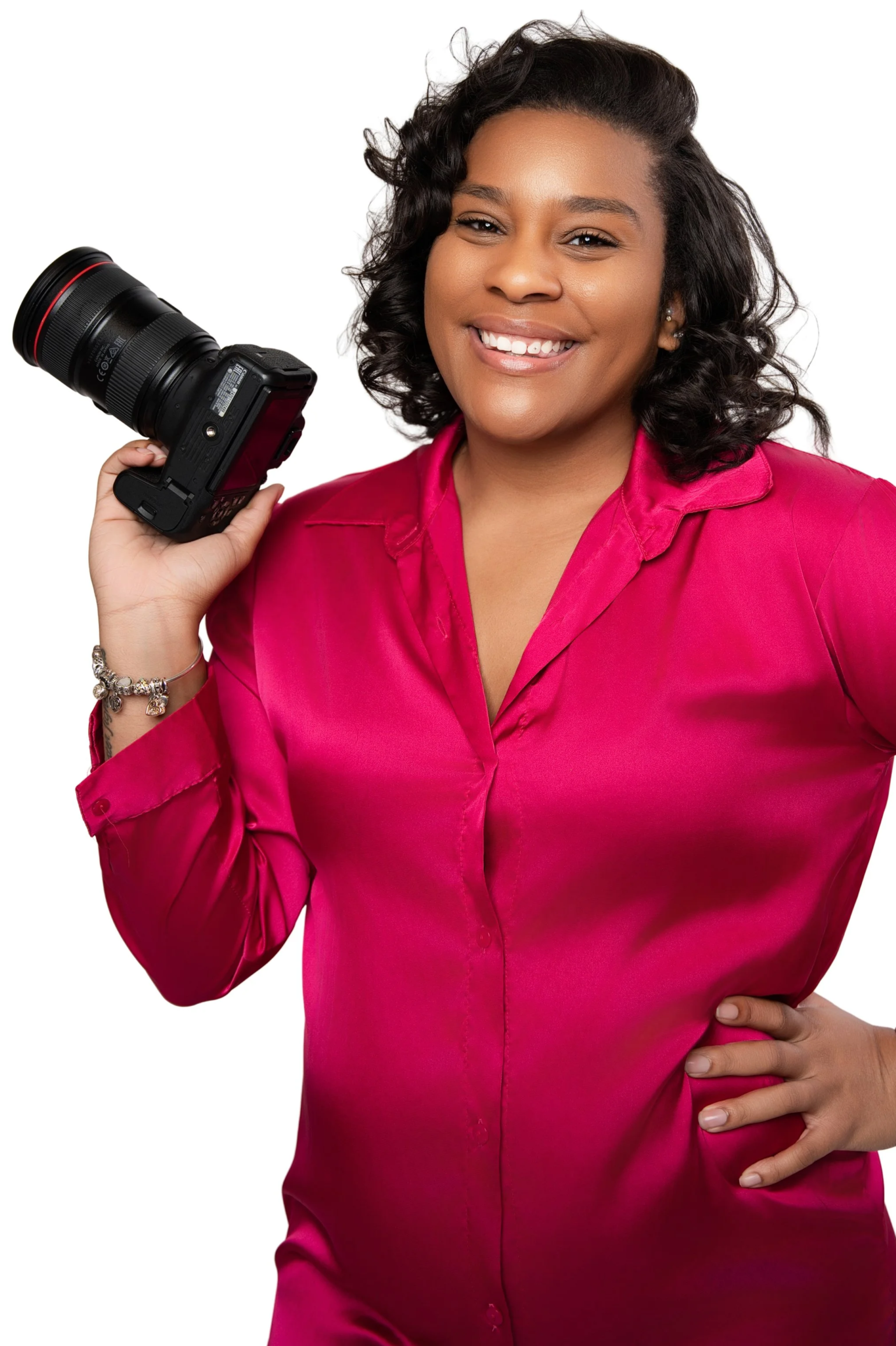 A woman smiling, wearing a pink satin blouse, holding a professional camera in her right hand against a white background.