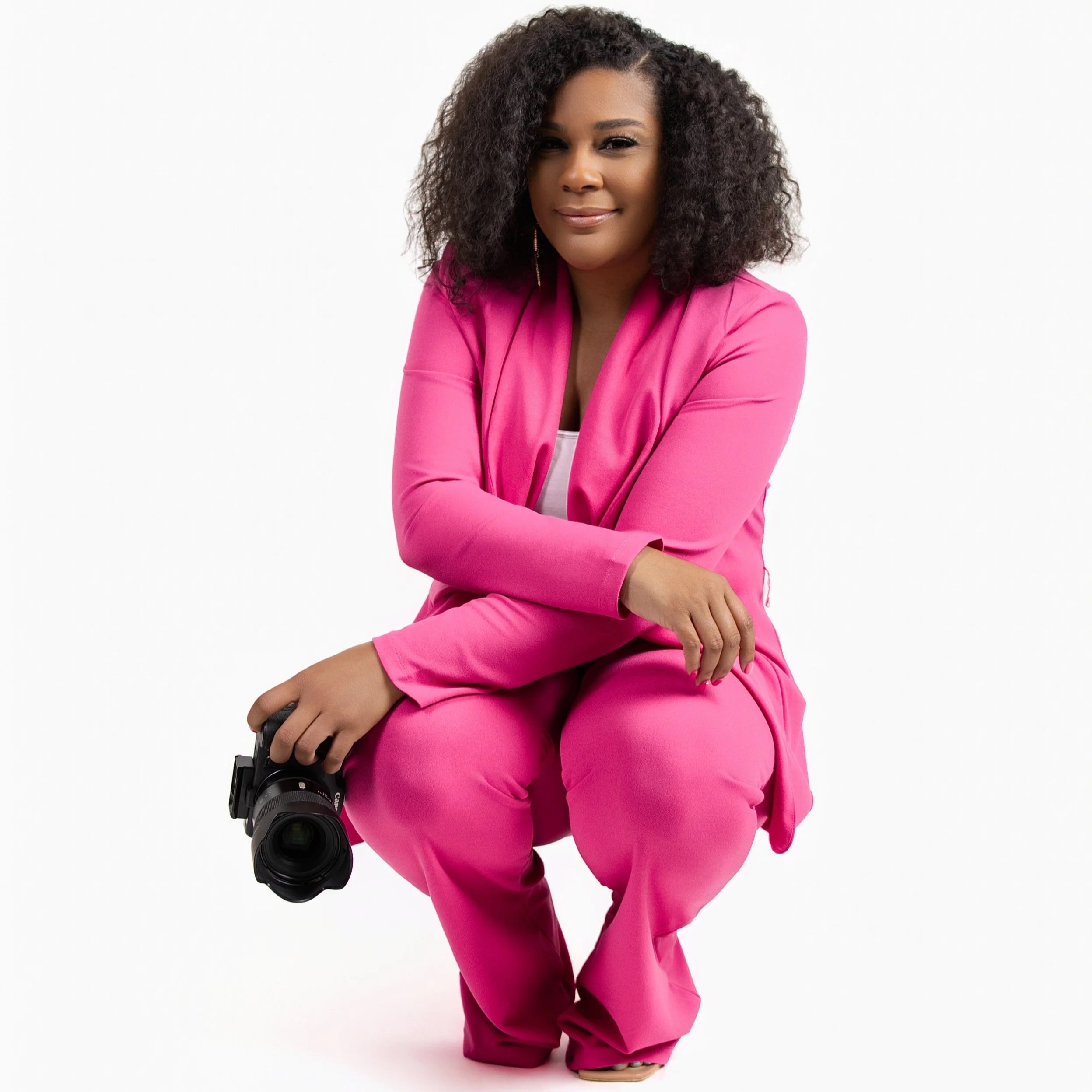 Woman with curly hair wearing a pink suit, squatting and holding a professional camera, against a plain white background.