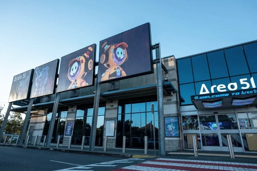 Exterior of Arena 51 with large computerized screens displaying cartoon astronauts and advertisements, located in a parking lot.