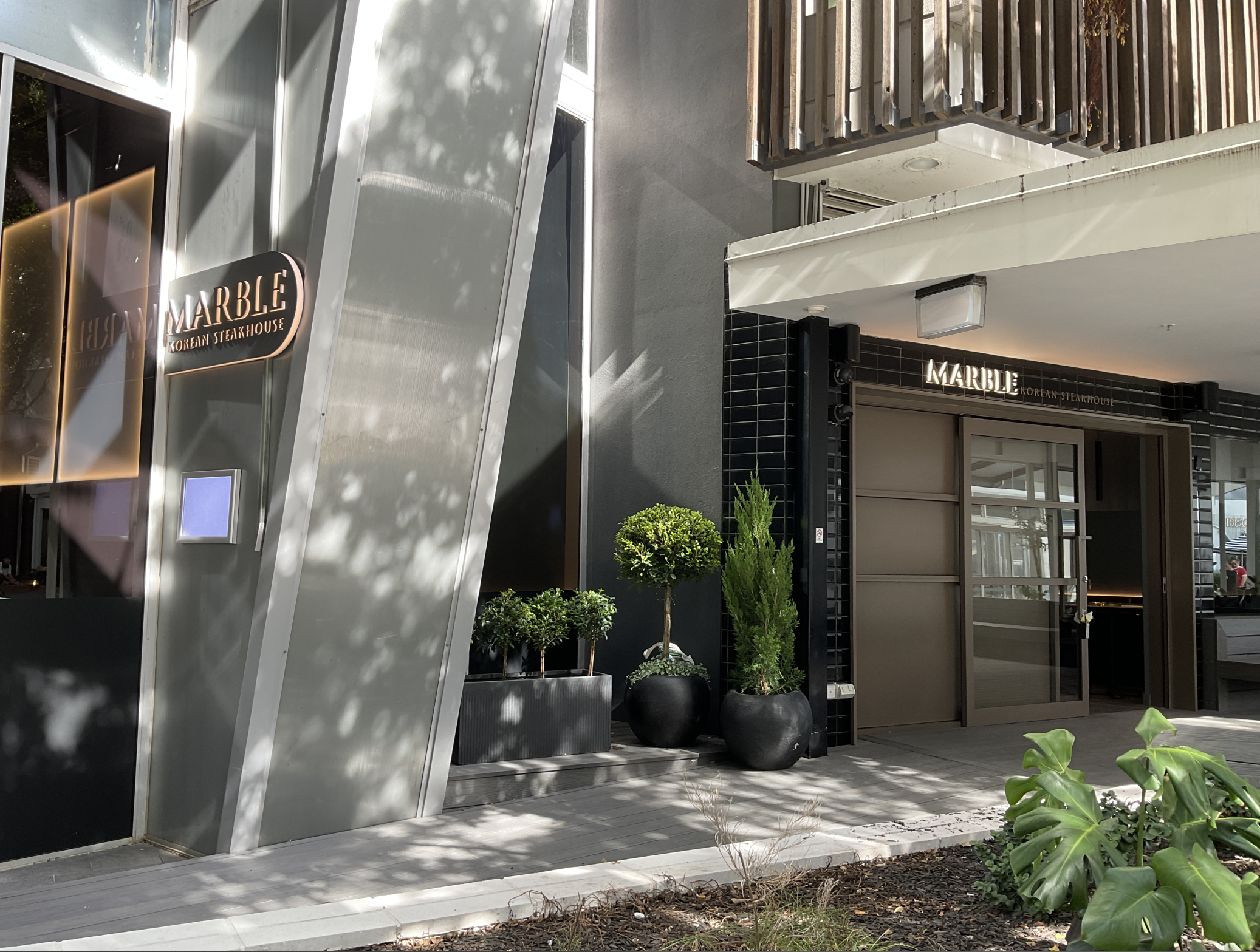Exterior view of Marble Korean Steakhouse with a modern entrance, black and beige color scheme, potted plants, and a sign above the door.