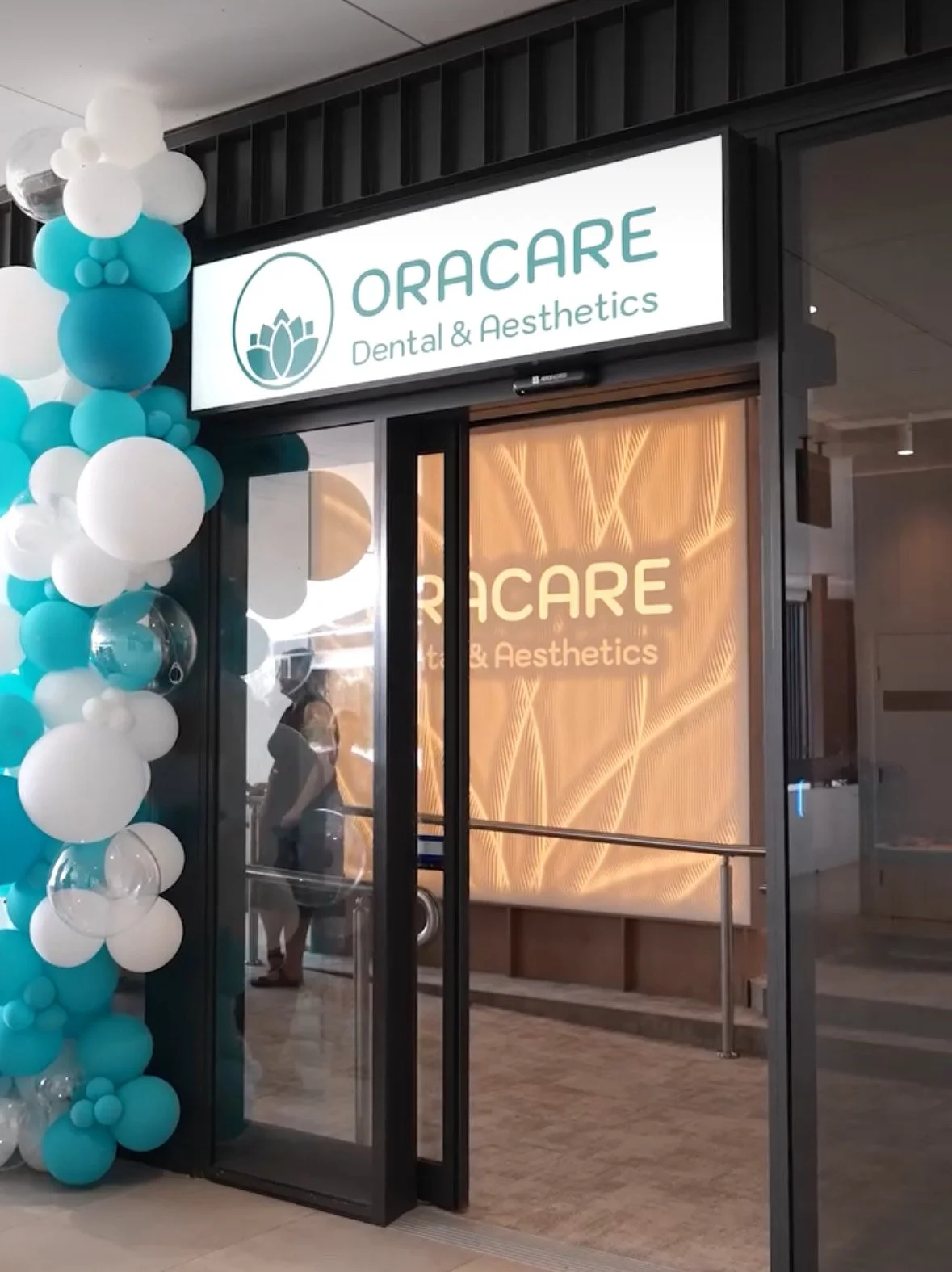 Entrance to ORACARE Dental & Aesthetics with glass doors, balloon decorations in white and turquoise on the side, and an illuminated sign above the door displaying the clinic's name and logo.