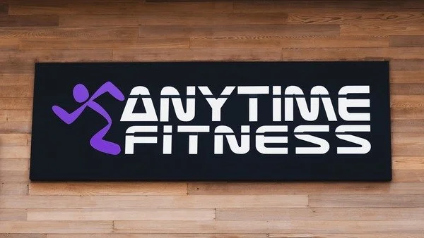 A black gym mat with a purple running figure and the words 'Anytime Fitness' in white on a wooden floor.