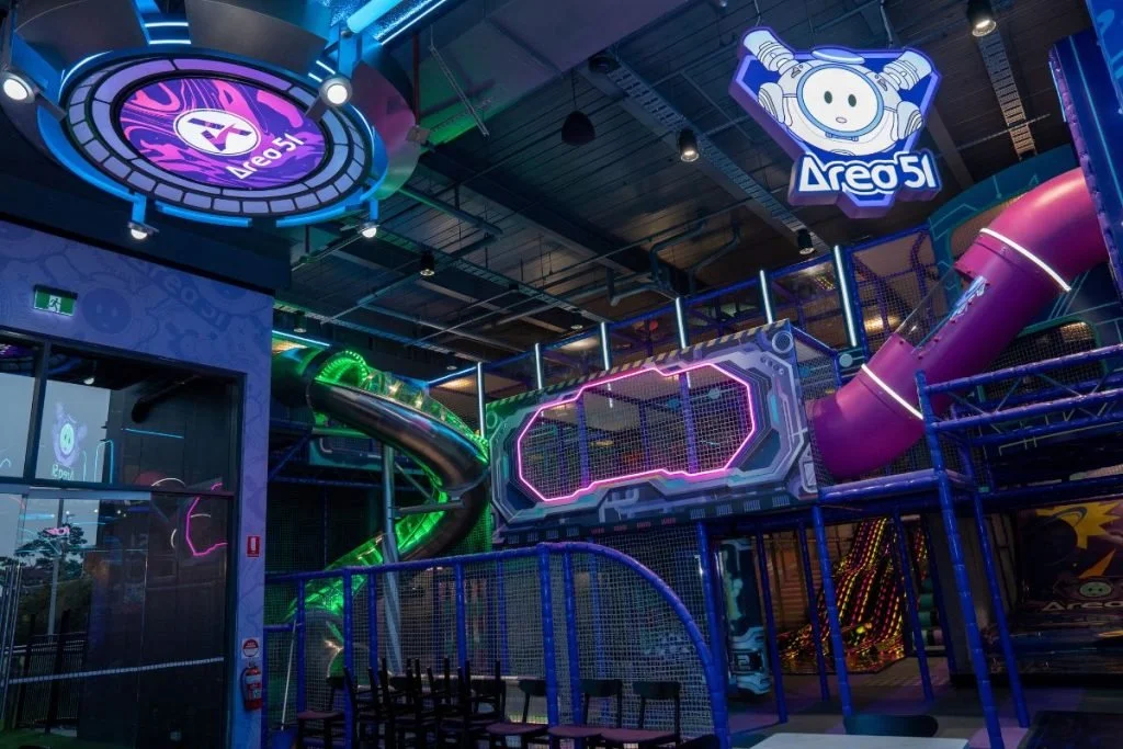 Indoor playground with colorful slides, a neon-lit maze, and signs with video game characters, including a circle face with headphones and a logo for 'Aeo 51'.