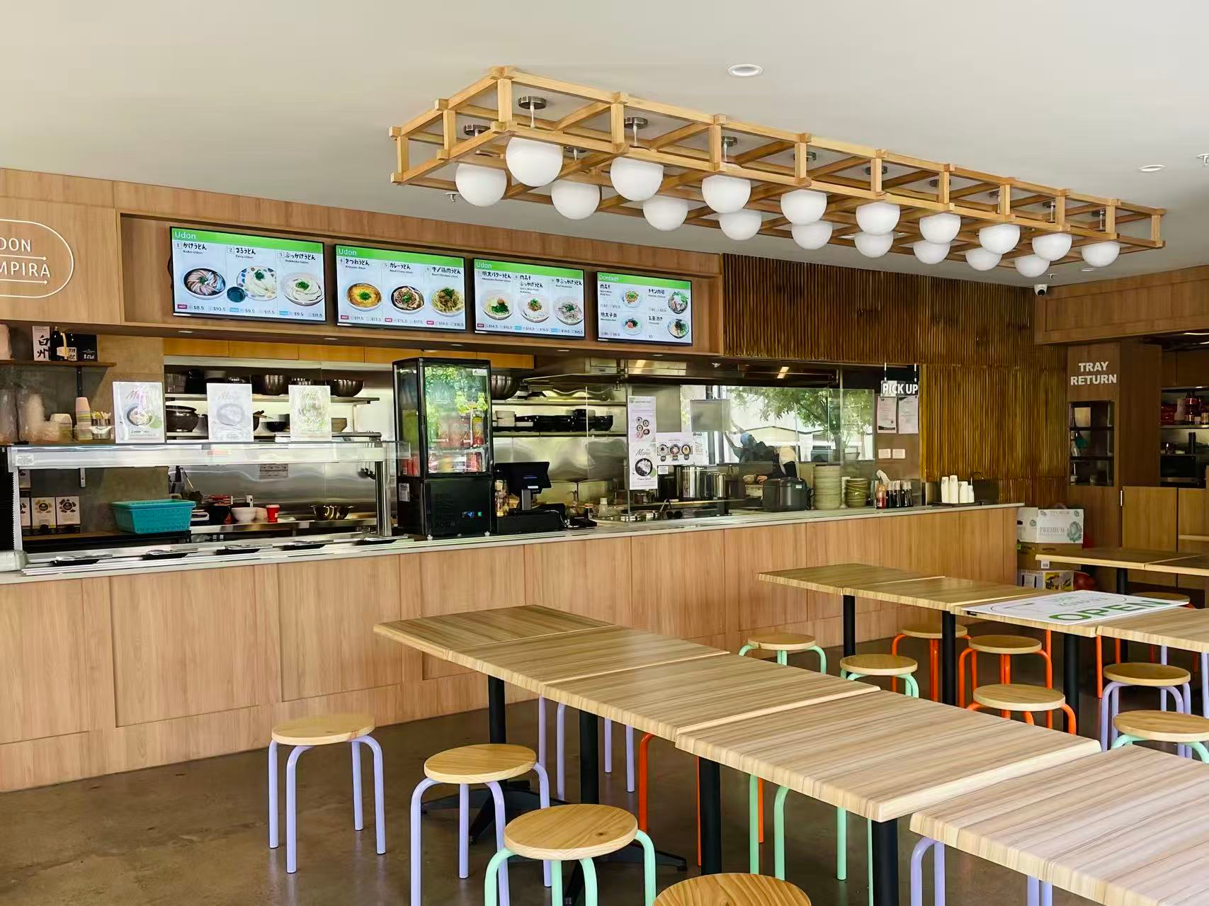 Empty restaurant with wooden tables, colorful stools, a counter with menu screens, and a kitchen visible in the background.