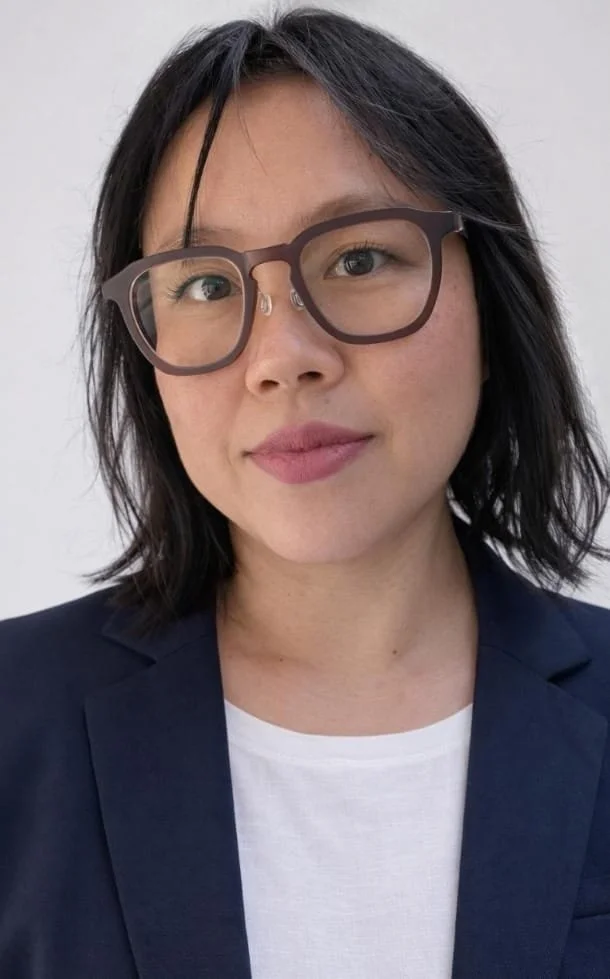 A woman with short black hair, wearing glasses, a dark blazer, and a white shirt, looking at the camera with a slight smile.