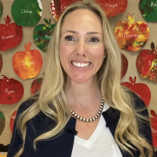 A smiling woman with long blonde hair wearing a pearl necklace and a black and white outfit, standing in front of a colorful school craft display of apples with children's names on them.
