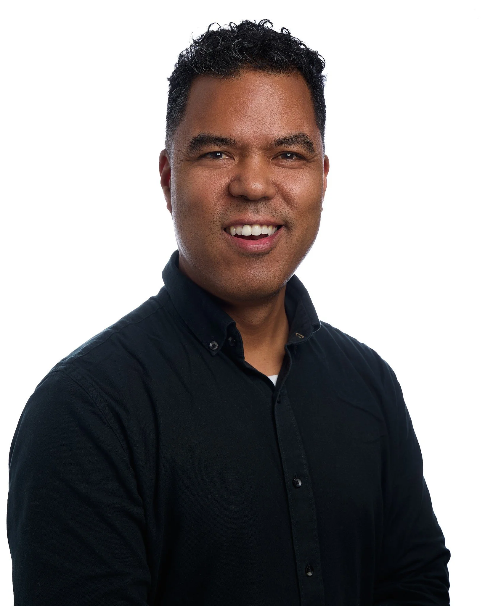 A man with curly black hair and a dark button-up shirt smiling at the camera against a plain white background.