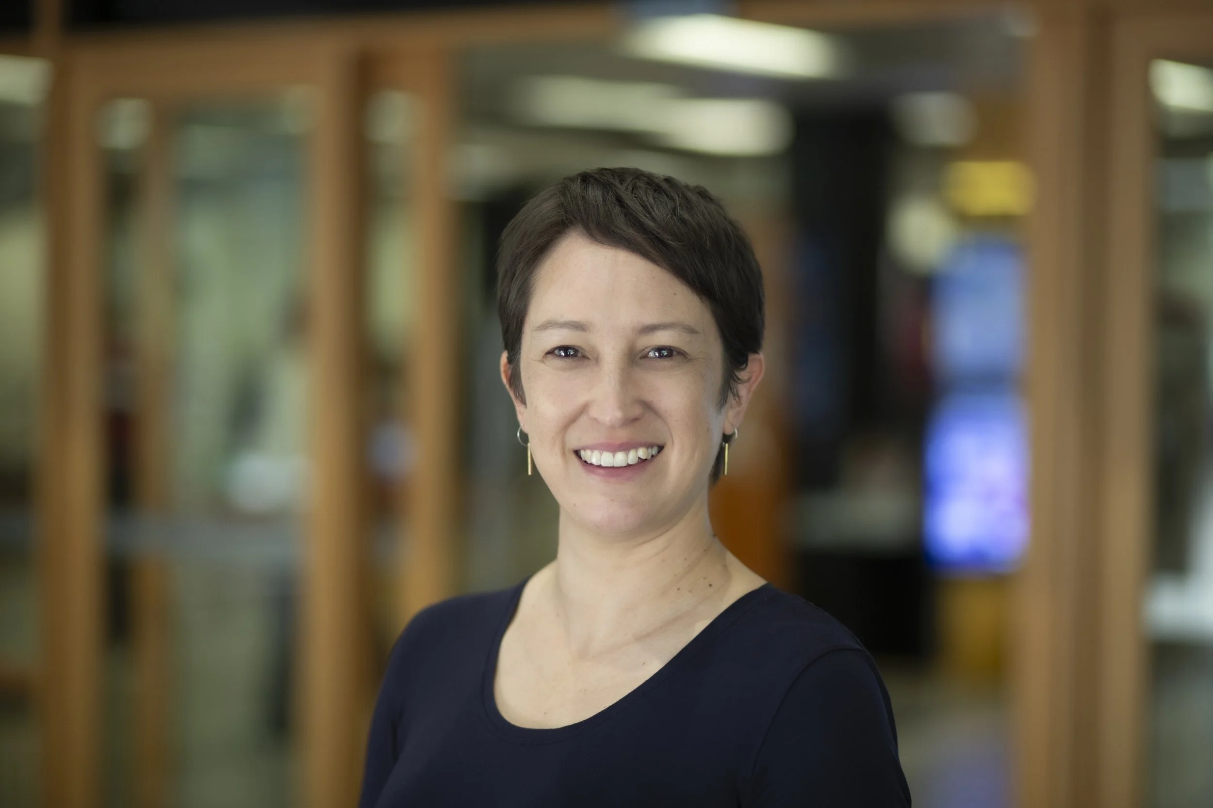 A woman with curly, shoulder-length brown hair and light skin, wearing a black pleated top with ruffled sleeves, standing against a plain white wall.