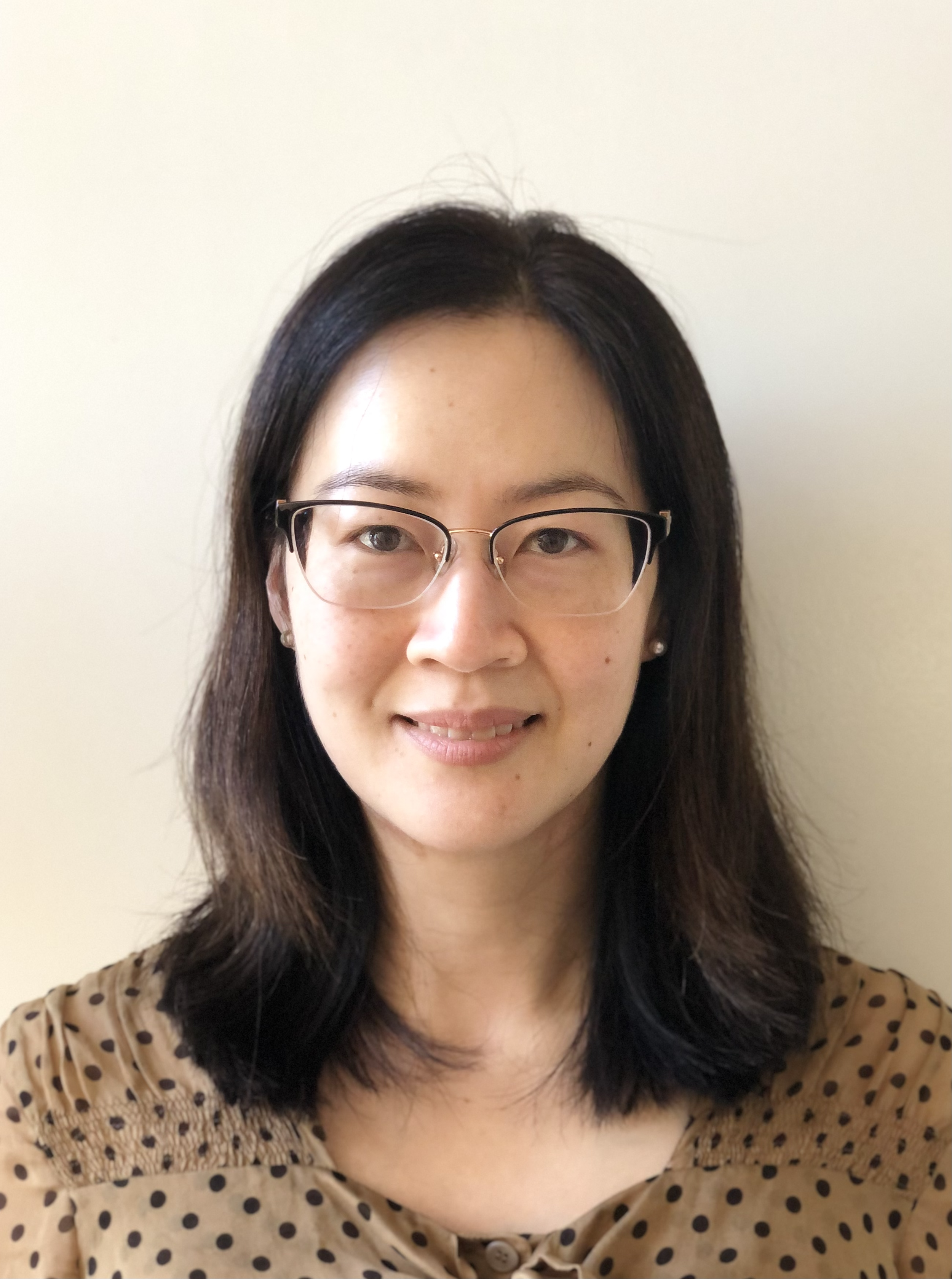 Close-up of a woman with black hair, glasses, and wearing a polka-dot blouse, smiling softly against a plain light-colored background.