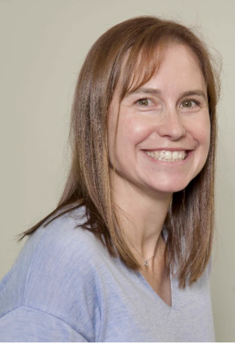 A woman with shoulder-length brown hair, smiling, wearing a light blue top, against a plain light-colored background.