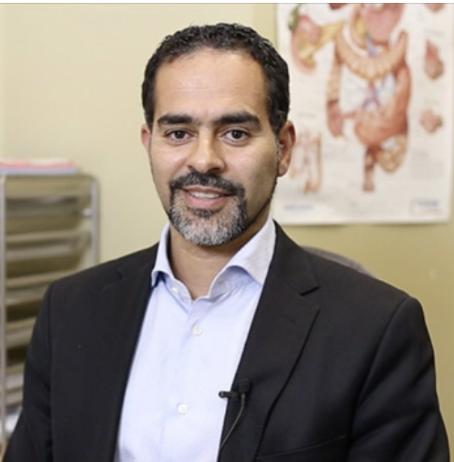 A man with dark hair, a beard, and mustache wearing a suit and light blue shirt, standing in an office with a medical chart in the background.