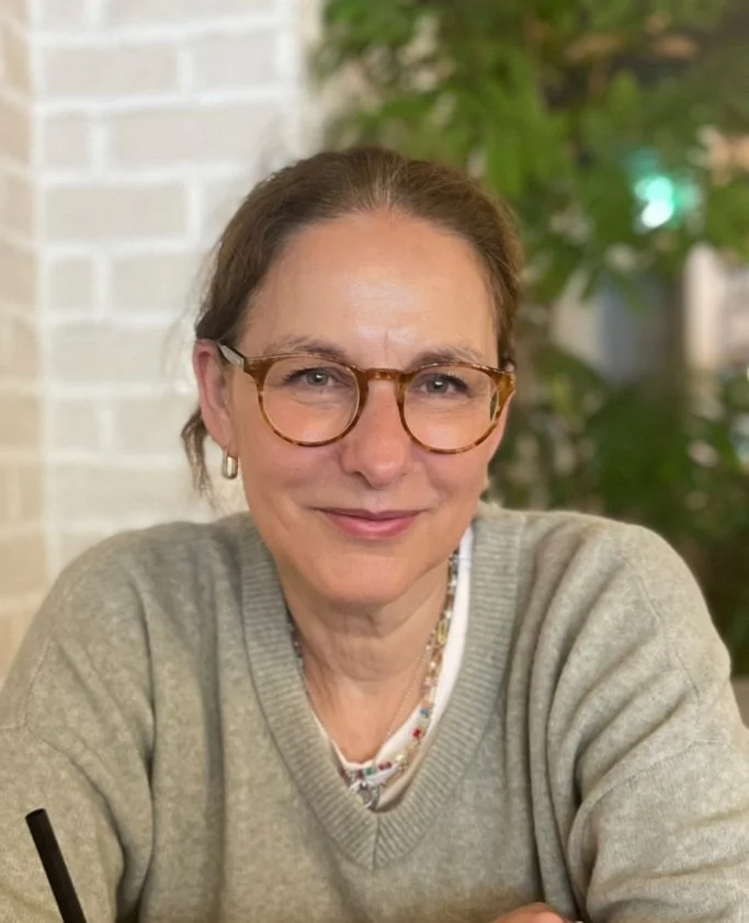 A middle-aged woman wearing glasses and a beige sweater, sitting indoors with a blurred green plant in the background.