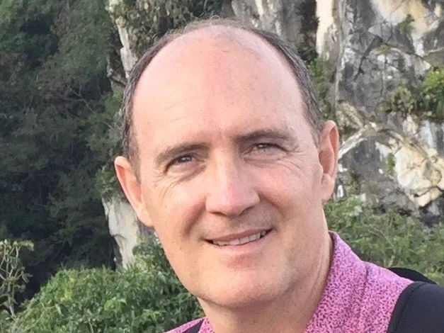 Close-up of a smiling man with short brown hair, light skin, wearing a purple shirt and carrying a black backpack, outdoors with trees and rocks in the background.