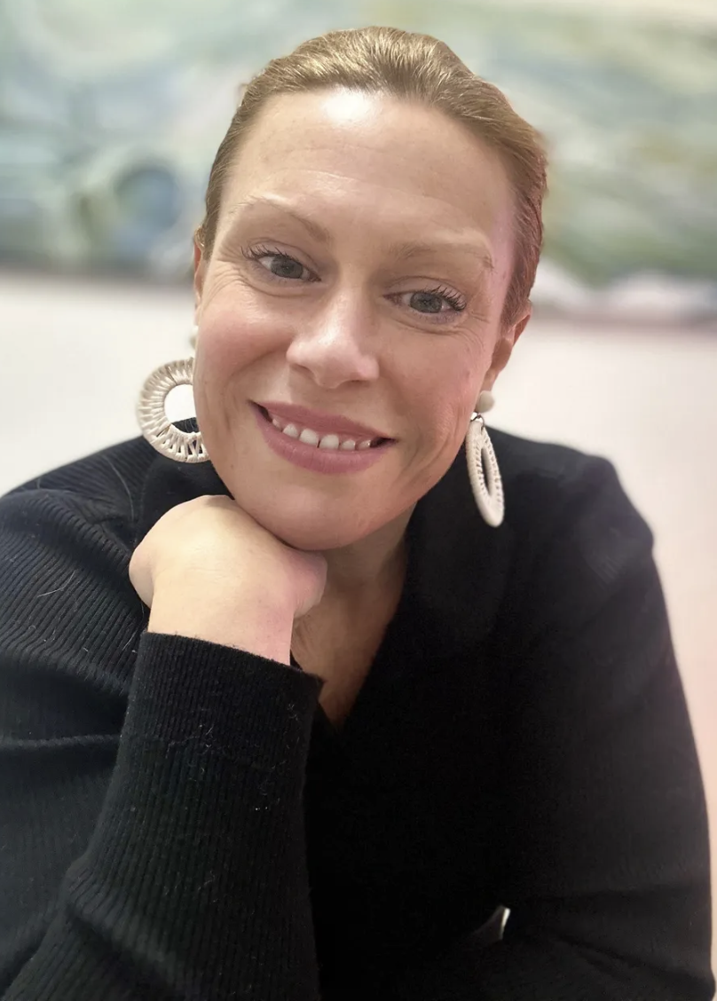 A close-up portrait of a smiling woman with fair skin, red hair pulled back, wearing large, circular earrings and a black top, resting her chin on her hand.