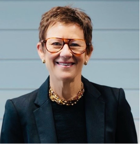 Smiling woman with short hair, glasses, gold jewelry, wearing a dark blazer, standing outdoors in front of a light blue wall.