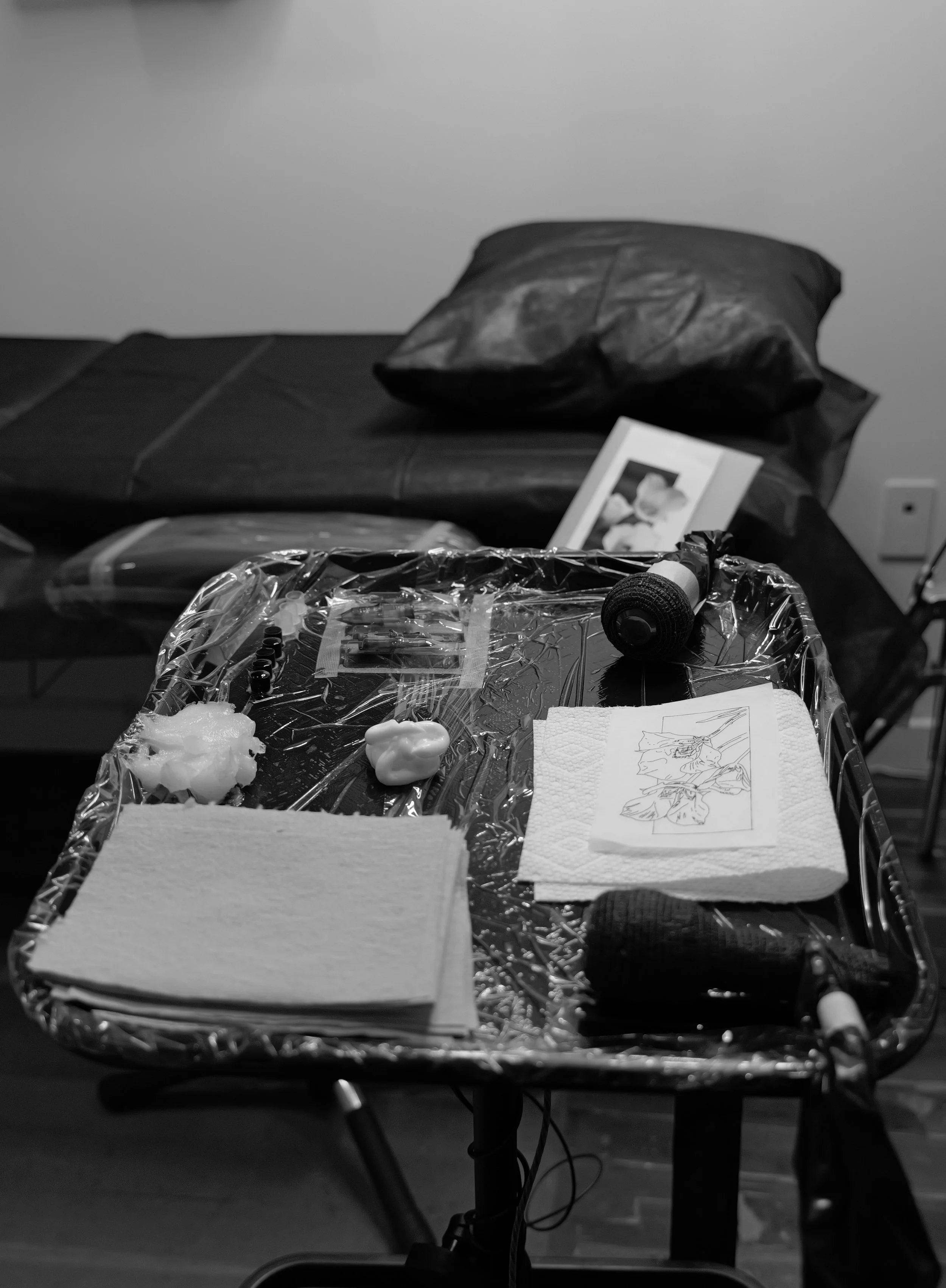 A black-and-white photo of a table with medical, sterile, clean tattoo supplies, including covered items, papers, tattoo machine, sterile tattoo needles, and a clean environment. A tattoo studio with comfortable bed and pillows for clients.
