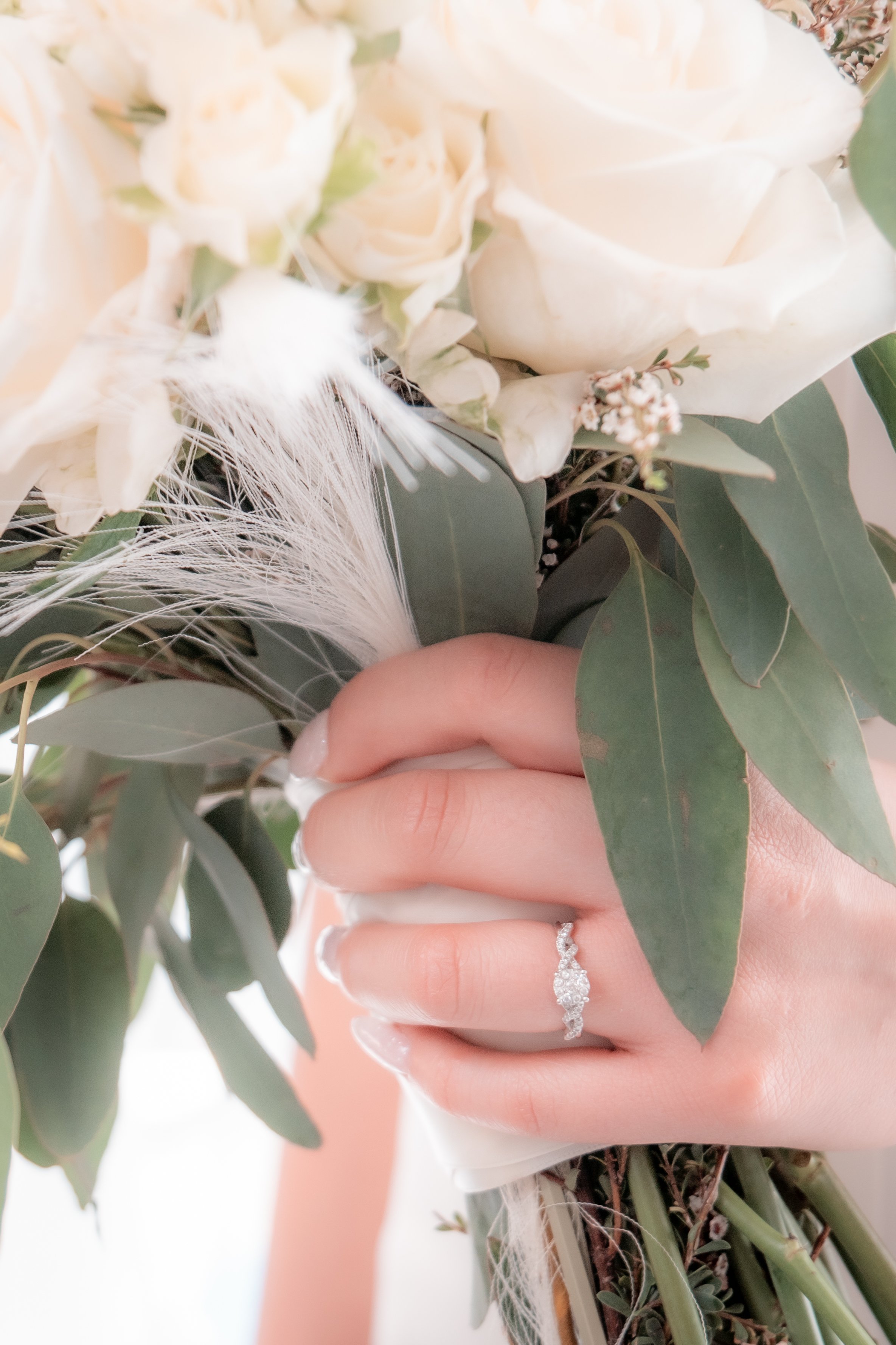 Close-up of a hand with an engagement ring holding a bouquet with white roses, eucalyptus leaves, and a white feather.