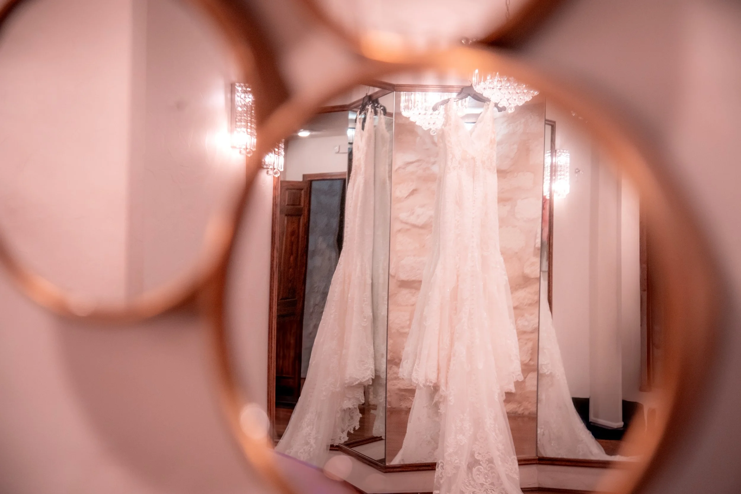 Wedding dresses hanging on a mirror in a boutique, framed by a decorative round mirror in the foreground.