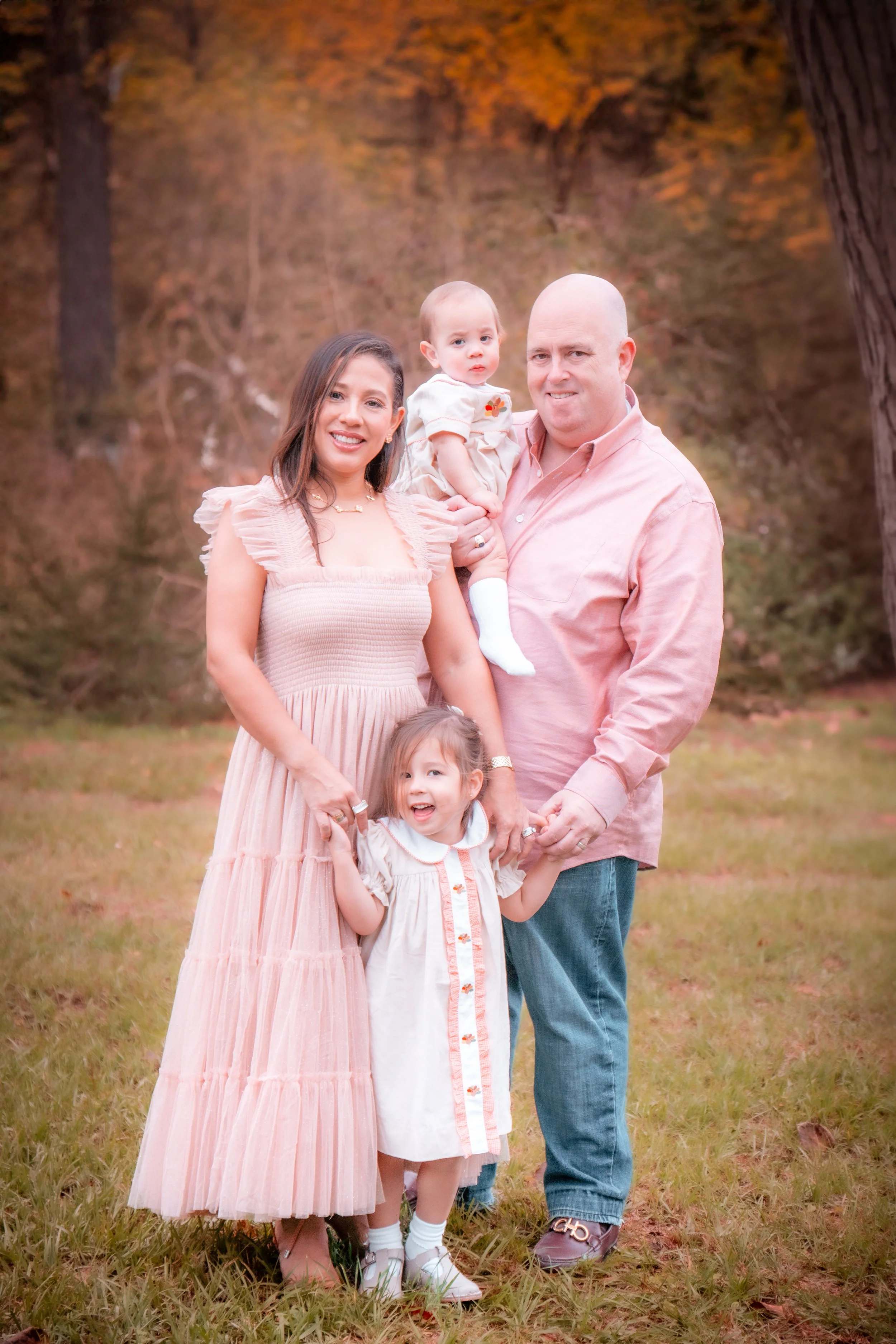 A family of four standing outdoors on grass, with fall foliage in the background. The mother has dark hair, wearing a pink dress. The father is bald, wearing a pink shirt and jeans. The daughter has light brown hair, wearing a white dress, and the ba