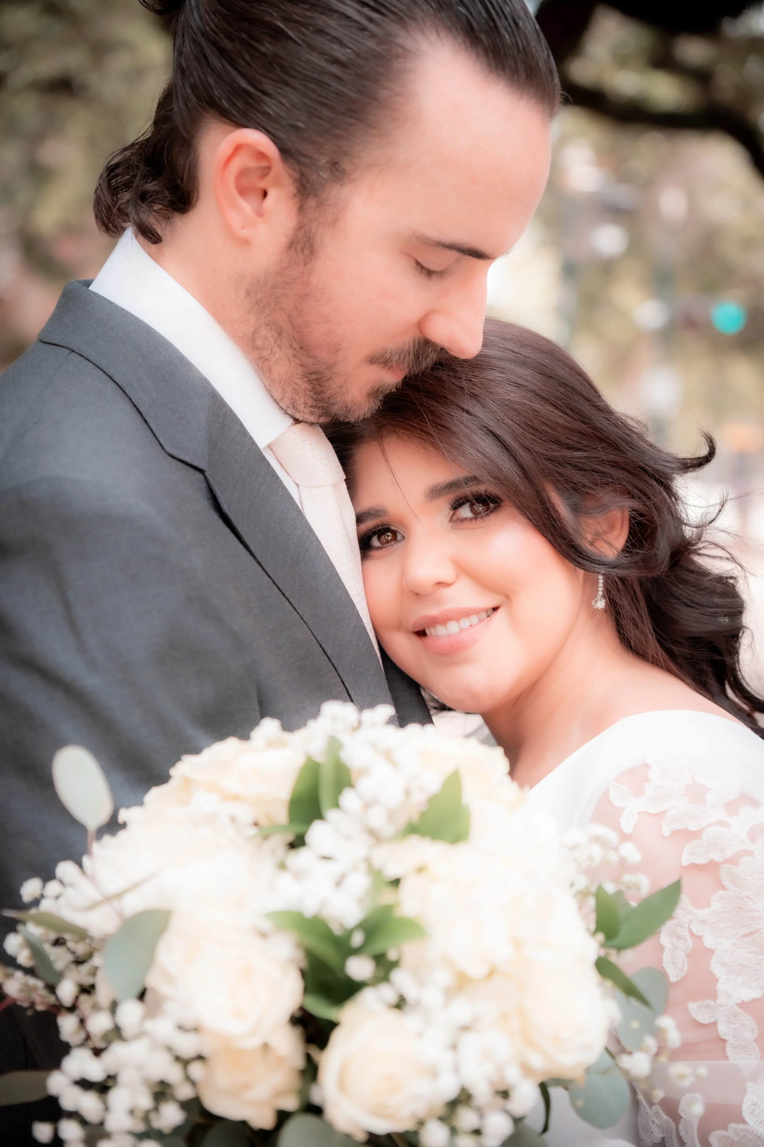 A bride and groom embrace outdoors, with the bride smiling at the camera and holding a bouquet of white roses and greenery.