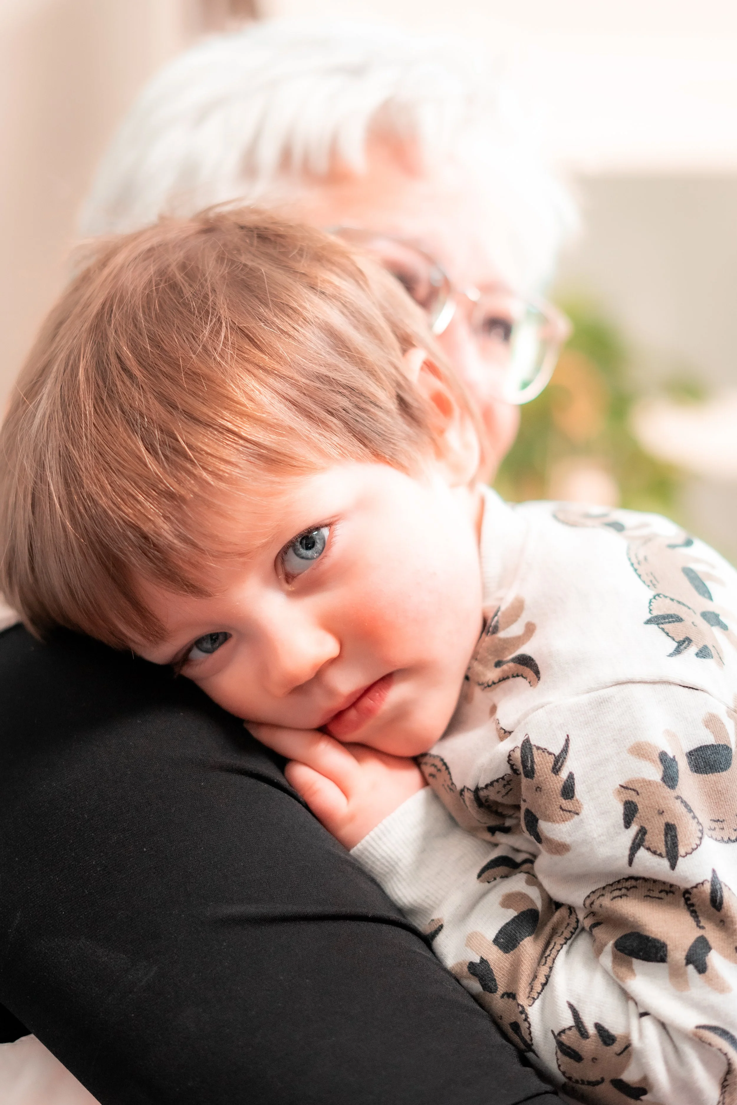 A young boy with blue eyes resting his head on an older woman's shoulder, appearing pensive and calm, in a close-up shot indoors.
