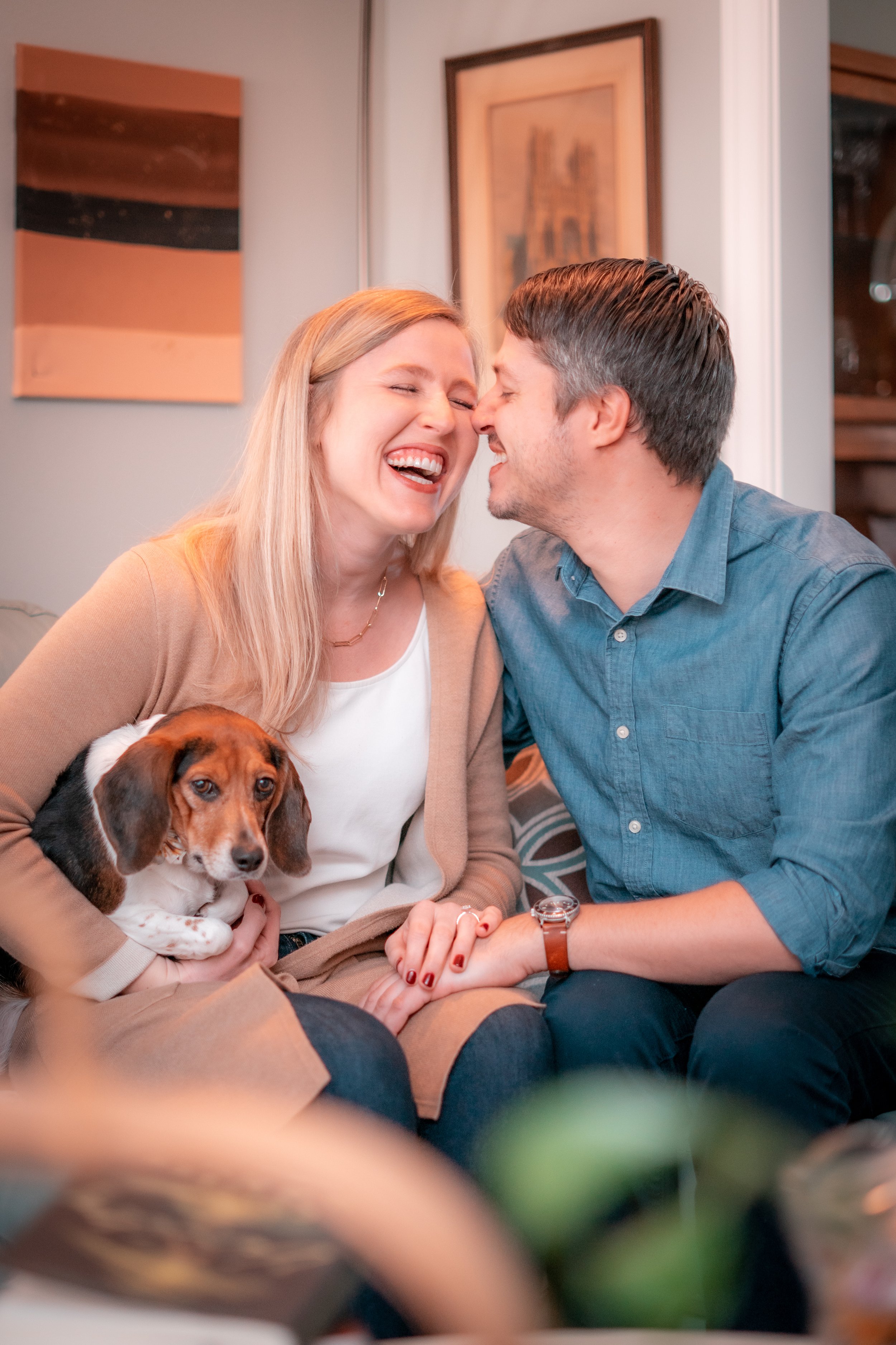 A couple sitting closely on a couch, smiling and touching noses, with a woman holding a small dog in her lap, in a cozy indoor setting with framed artwork on the wall.