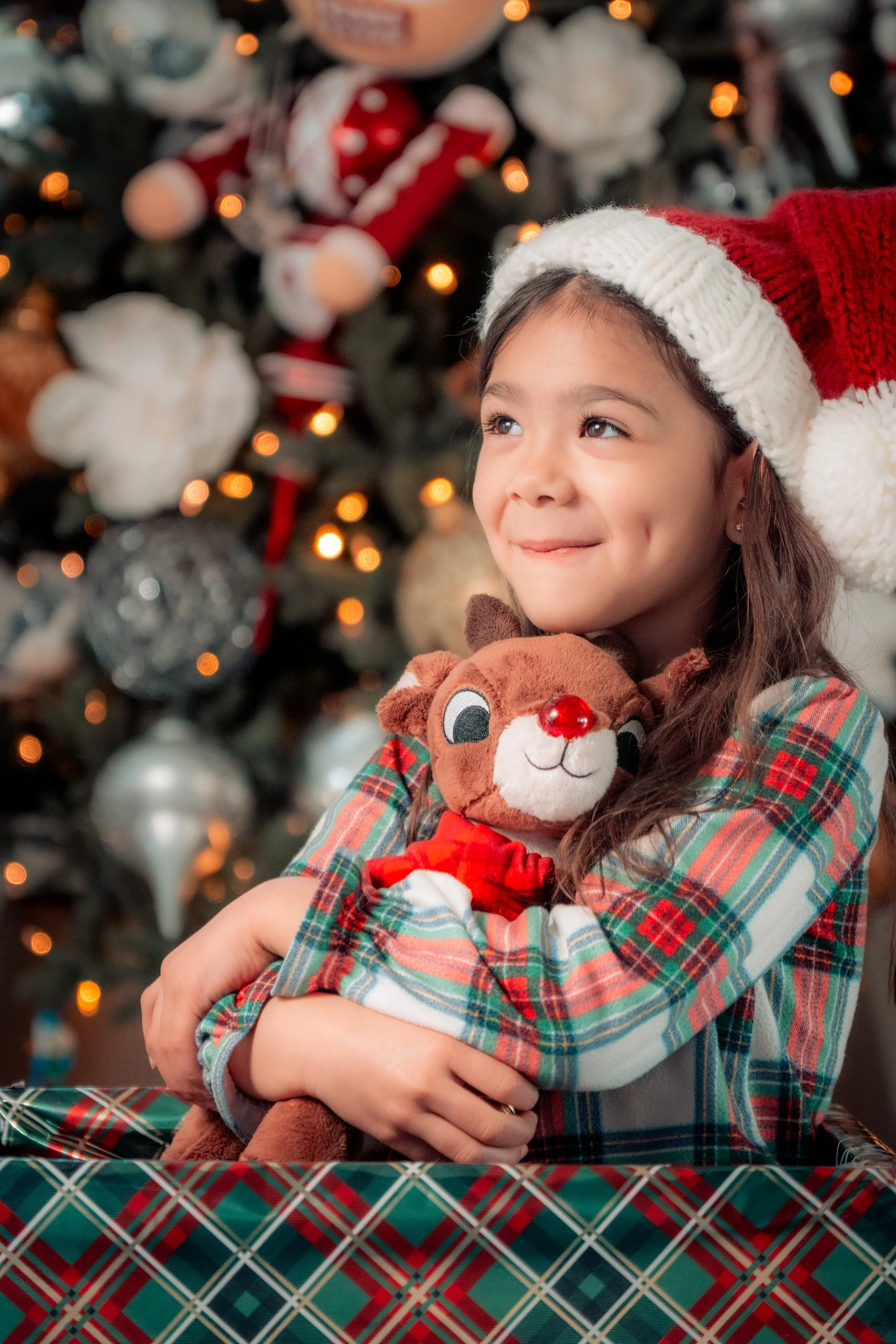 A smiling young girl wearing a Santa hat, hugging a plush reindeer toy, with a decorated Christmas tree in the background.