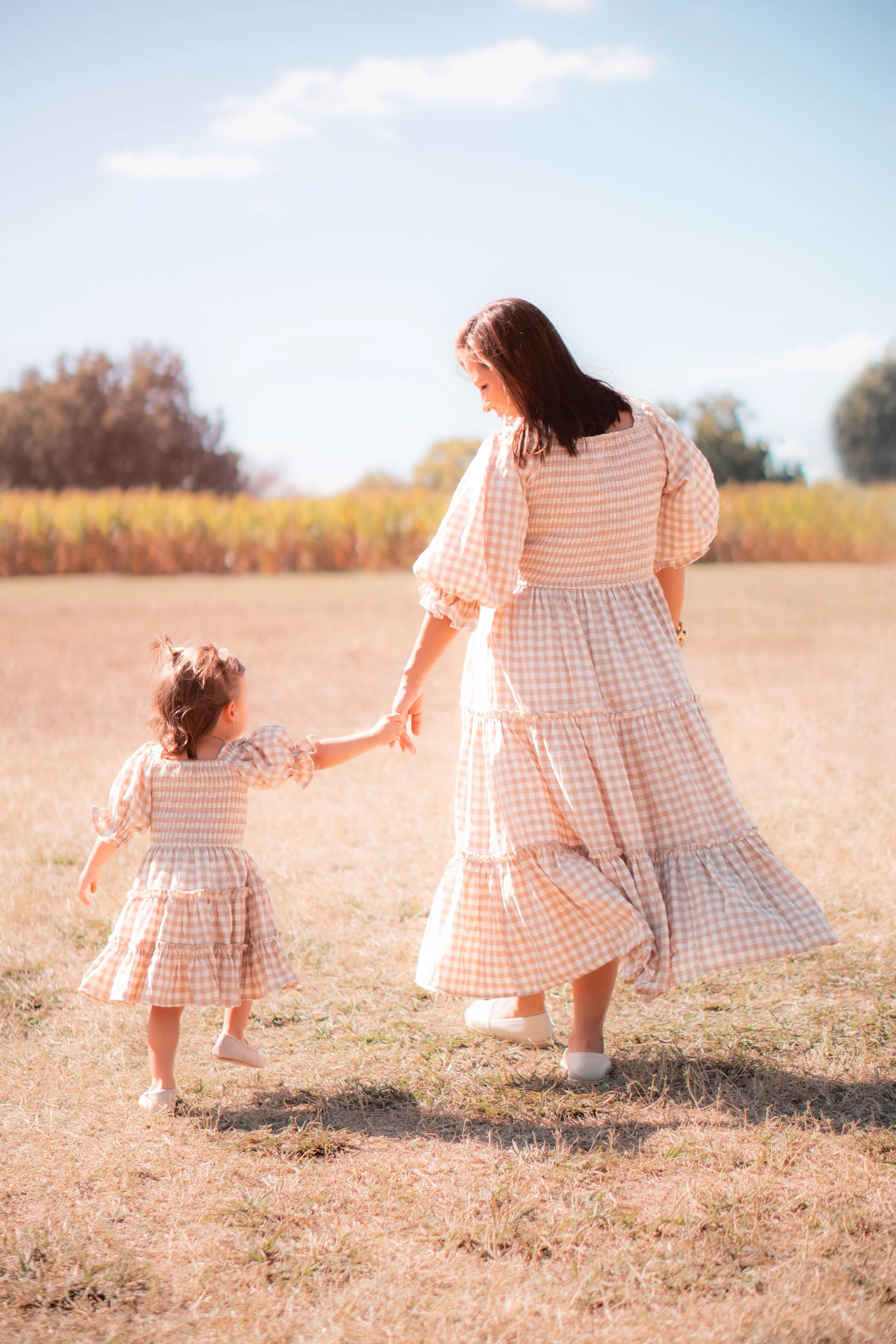 A woman and a young girl holding hands and walking in a field on a sunny day, both wearing matching beige and white gingham dresses.