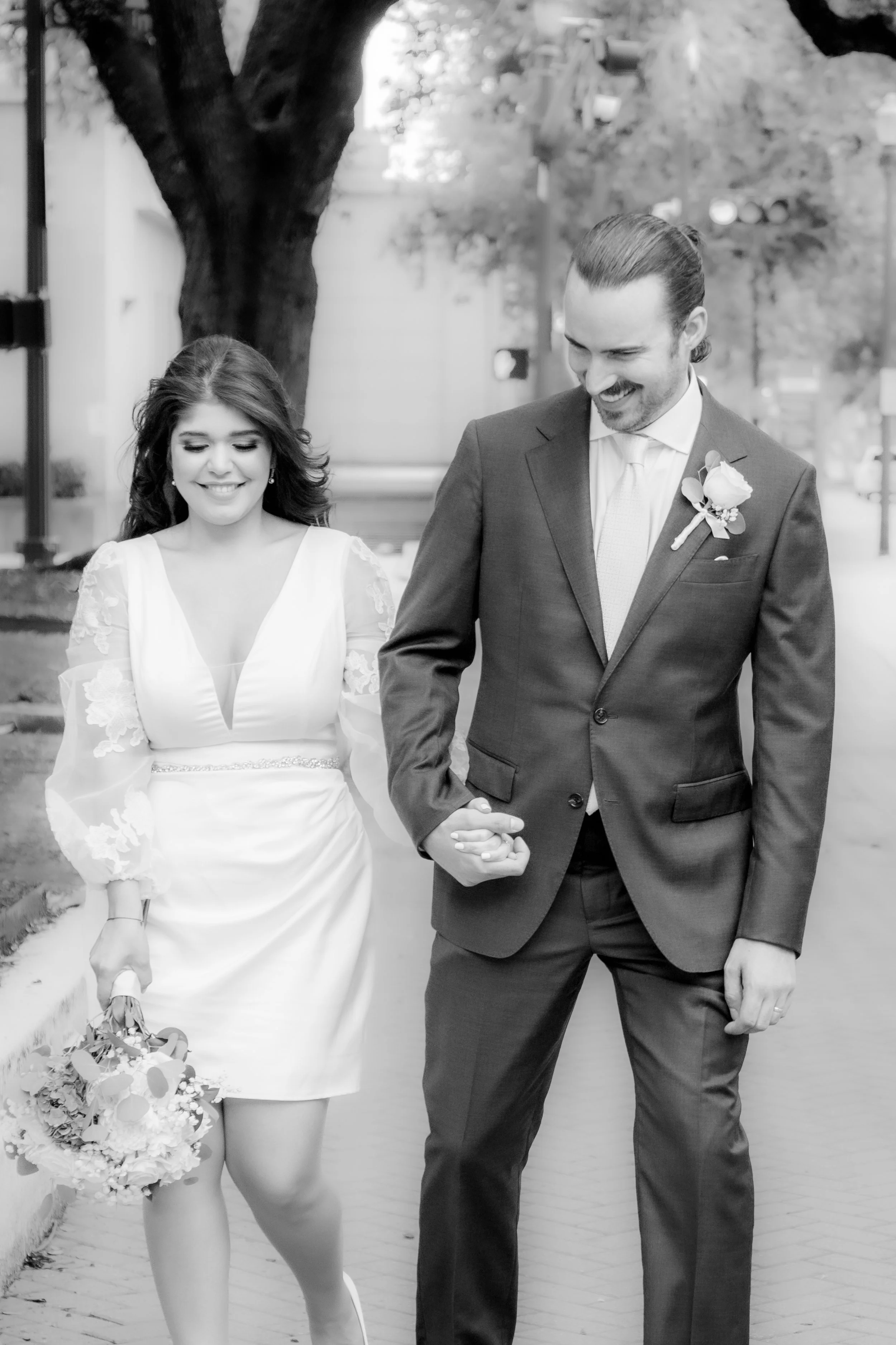 A happy couple in wedding attire walking outdoors, holding hands and smiling, with a large tree and urban background.