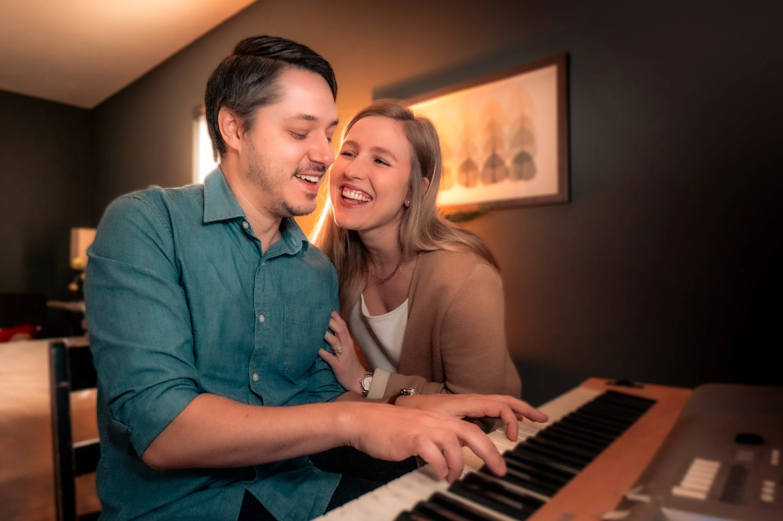 A young couple joyfully playing piano together in a dimly lit living room, smiling with affection.