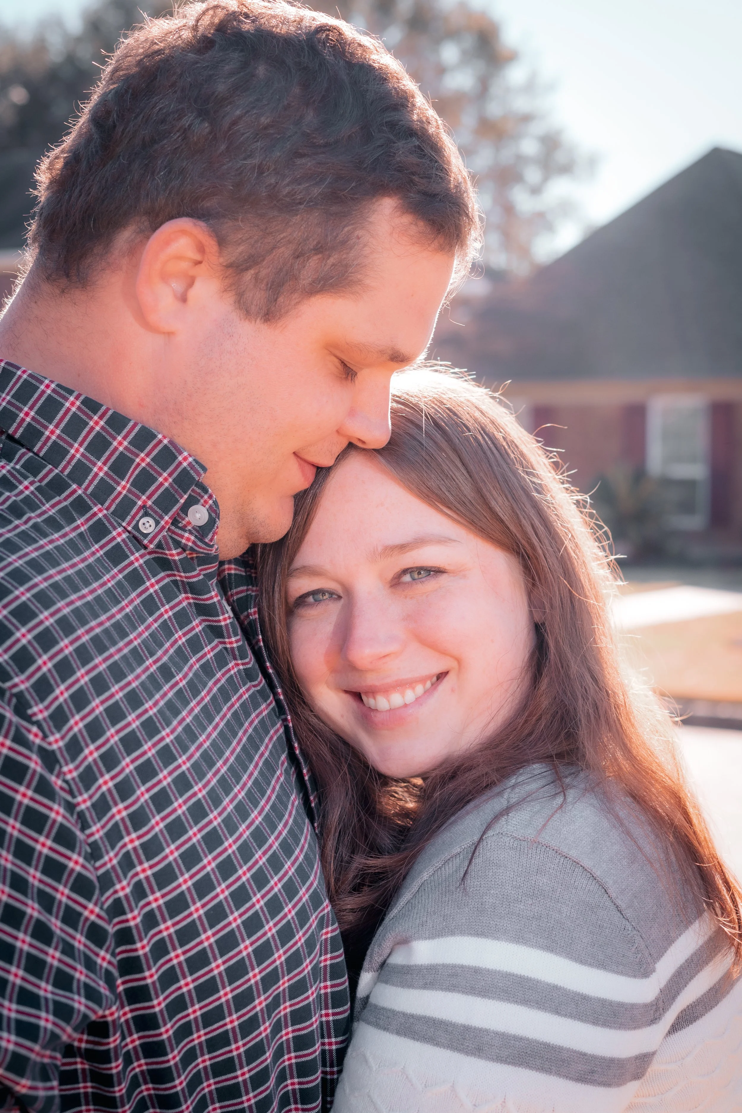 A young couple sharing an intimate moment outside on a sunny day, with the man gently touching his forehead to the woman's head and both smiling.