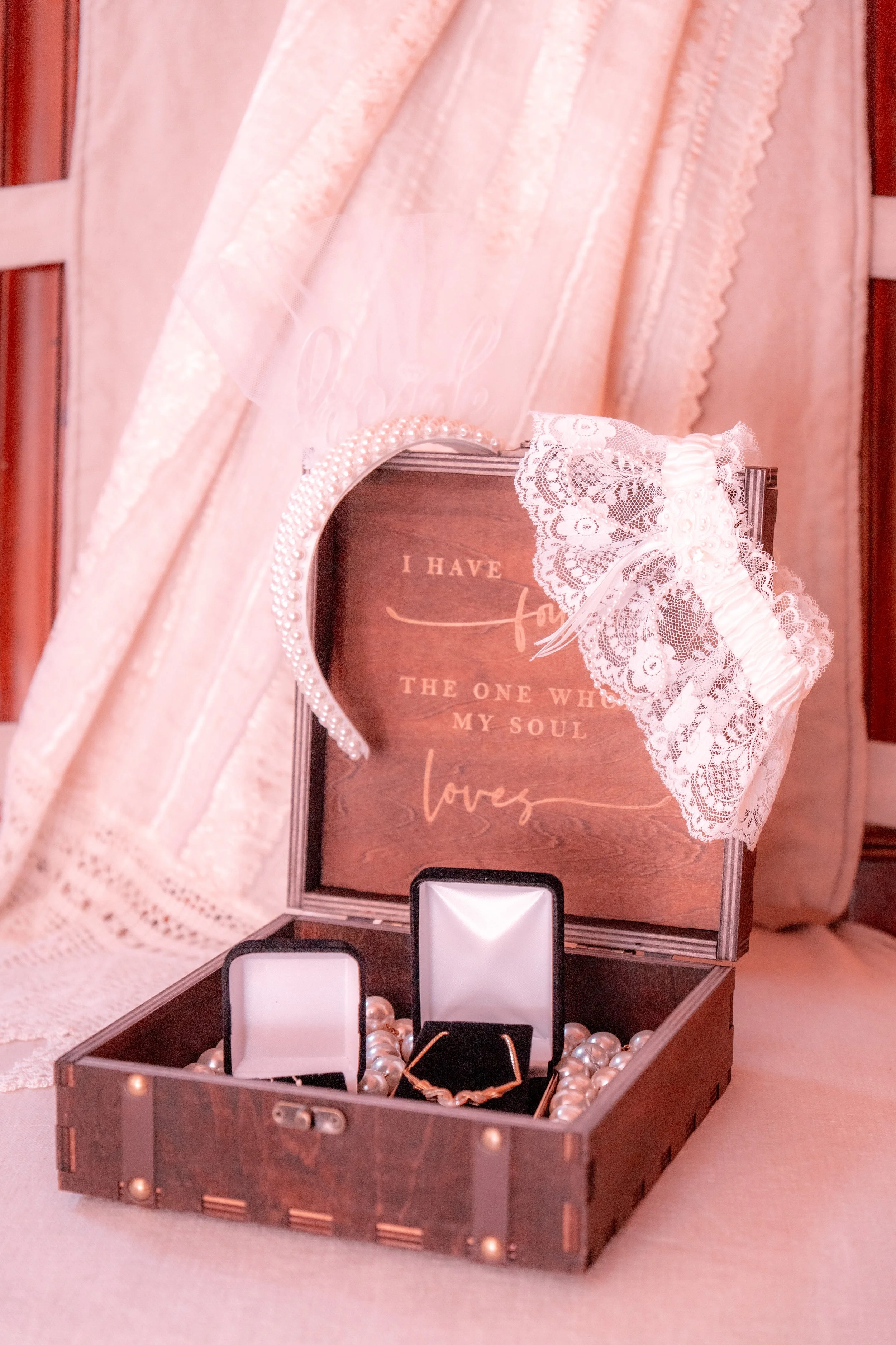 A wooden jewelry box containing pearl necklaces, earrings, and a bracelet, with lace and pearl accessories draped over the open lid. Behind the jewelry box, a framed quote reads 'I have found the one whom my soul loves,' with a pink lace ribbon bow a